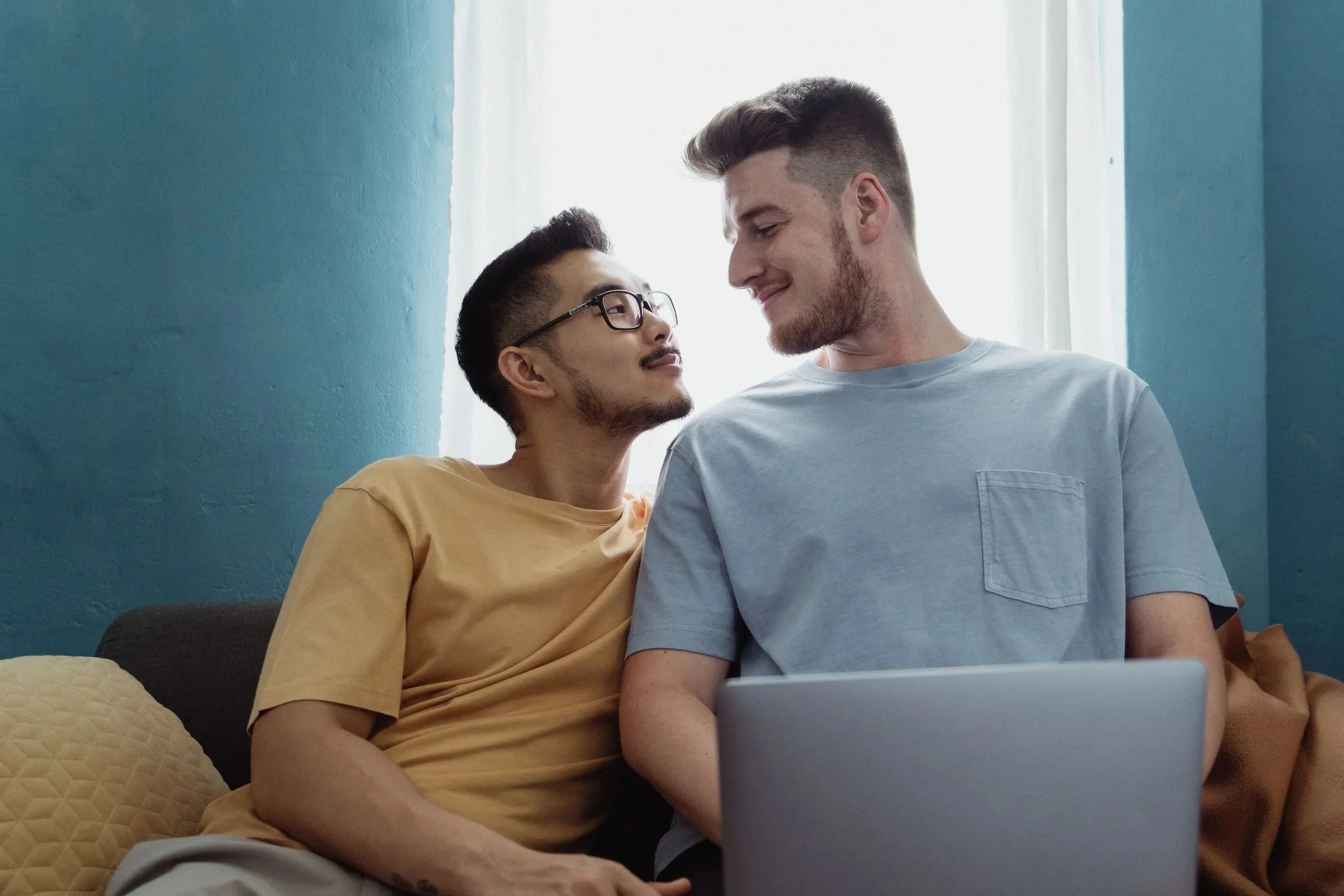 Two young men sitting close together, smiling, with laptops in front of them, in a room with teal walls and a white curtain.