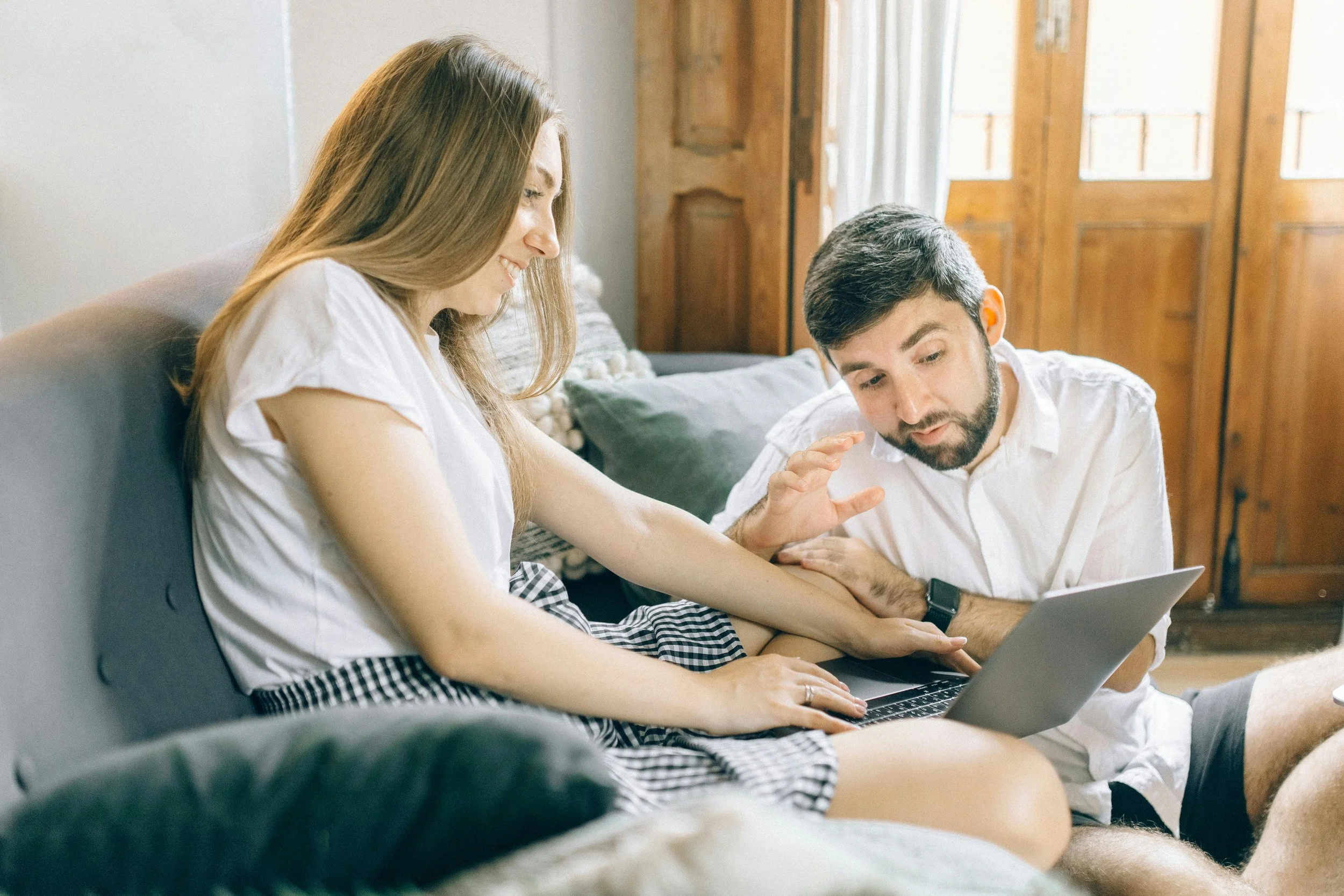 A woman and a man sitting on a couch, looking at a laptop together, smiling and engaging with each other in a cozy room with wooden accents.