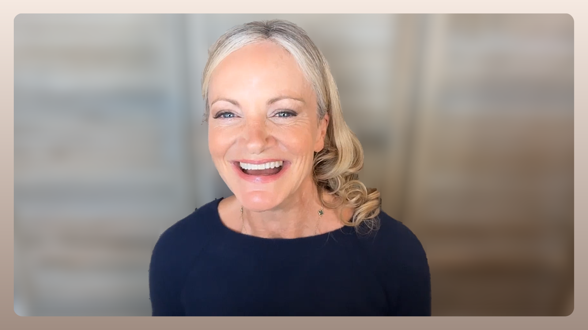 A middle-aged woman with blonde hair styled in loose curls, smiling warmly and looking directly at the camera, wearing a black top and a delicate necklace, with a softly blurred background.