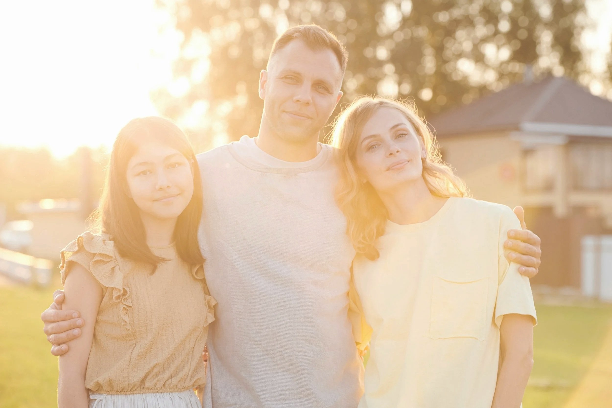 A group of three people, consisting of a man and two women, standing closely together outdoors during sunset or sunrise, smiling softly, with trees and a house in the background.
