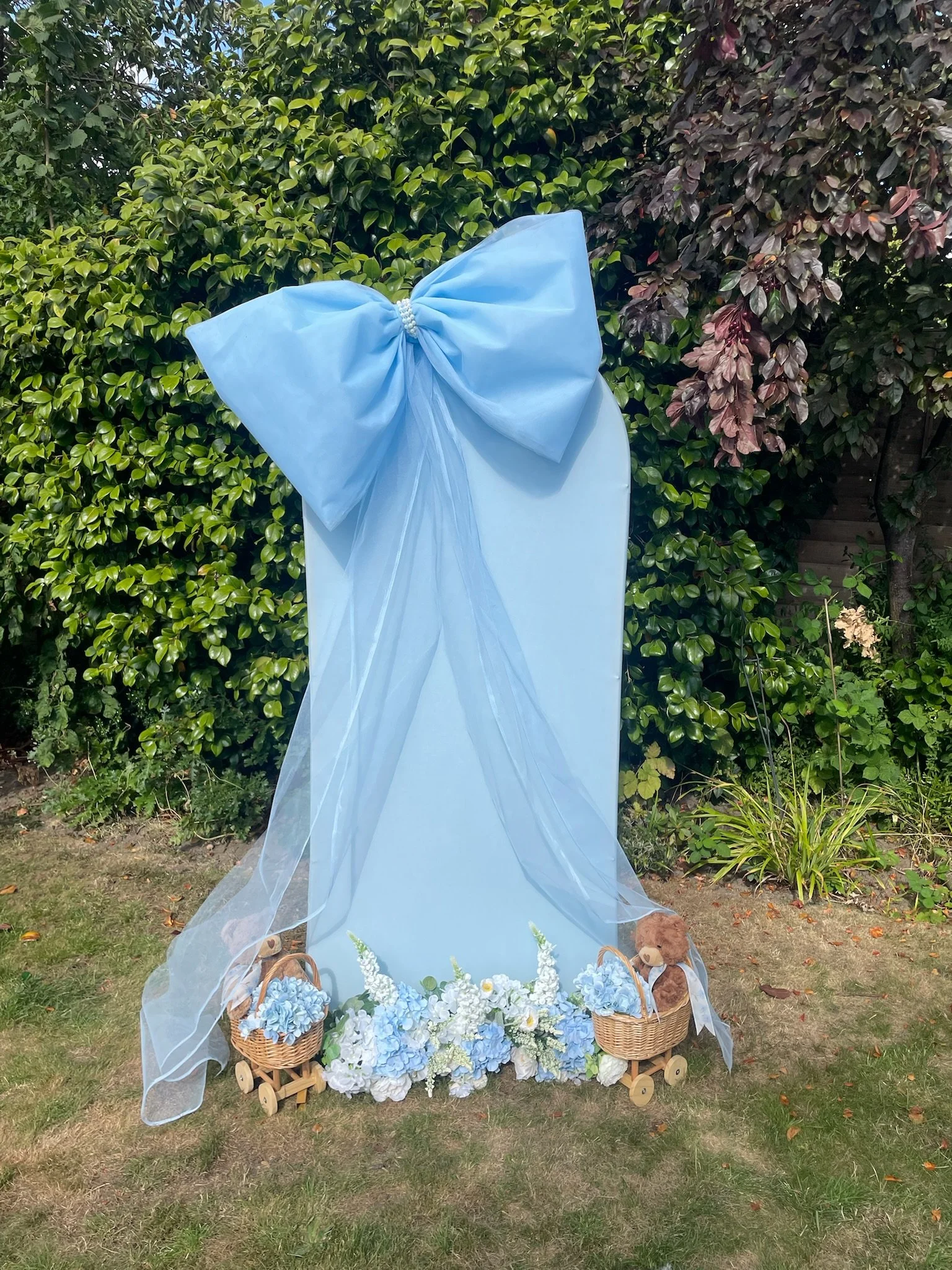 A decorative outdoor display featuring a large blue bow and sheer fabric draped over a white panel, flanked by baskets of blue and white flowers and small teddy bears on a lawn with bushes and trees in the background.