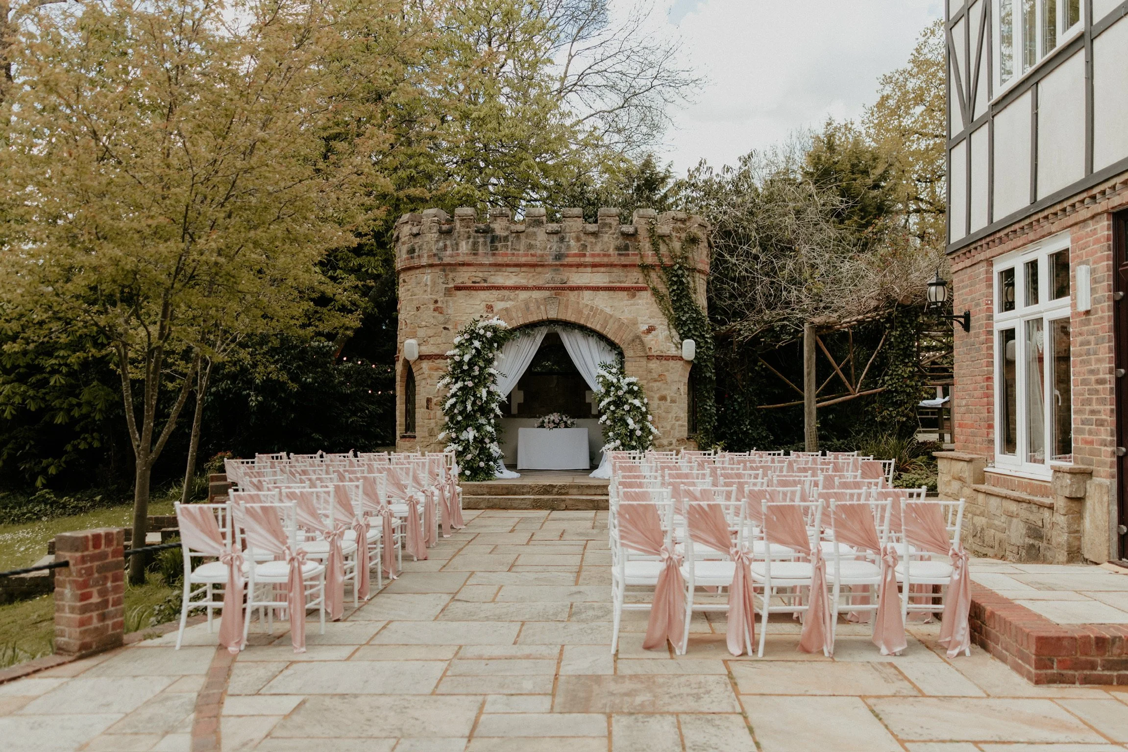 Outdoor wedding setup with rows of white chairs decorated with pink ribbons facing a small brick chapel with floral arch, set in a garden with trees and a stone building on the right.
