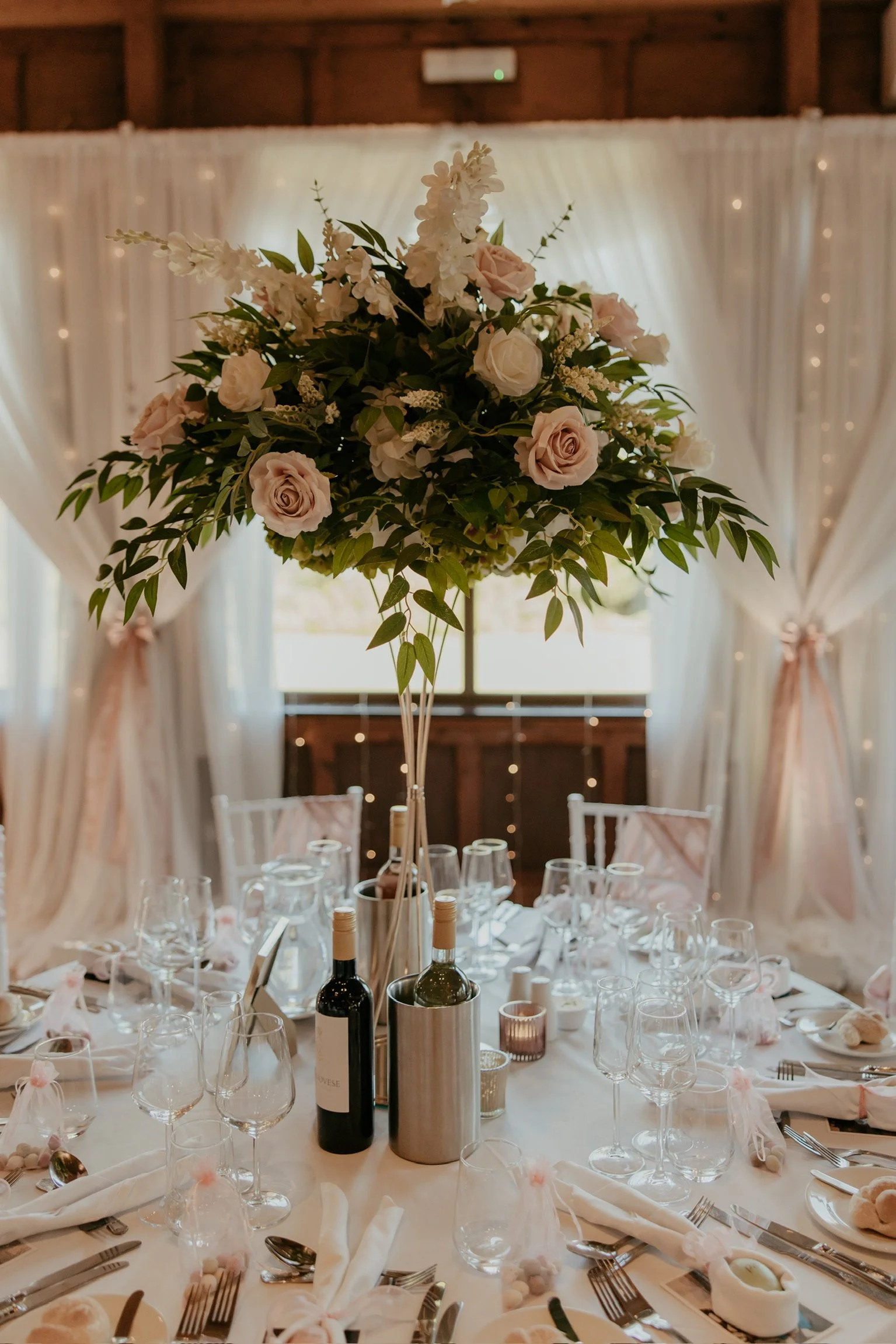 Elegant wedding reception table with a tall floral centerpiece of roses and greenery, surrounded by wine glasses, plates, utensils, and decorated with pink ribbons and small pom-poms, with white draped curtains and string lights in the background.