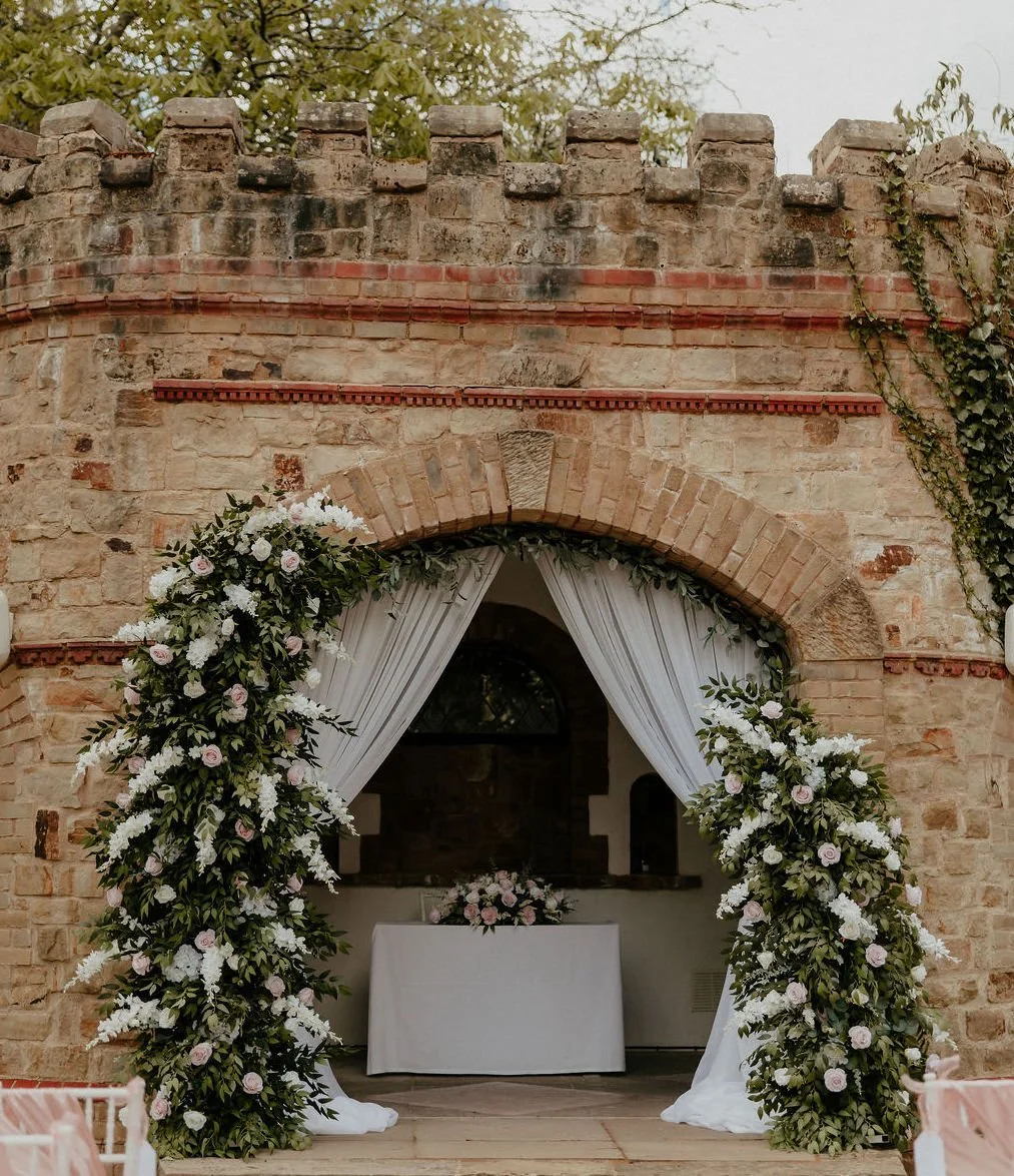 A rustic stone archway decorated with a lush floral arch of pink roses, white flowers, and greenery. With top table white and pink roses, daisy's and greenery, florals which can be Ultalized for ceremony and wedding breakfast
