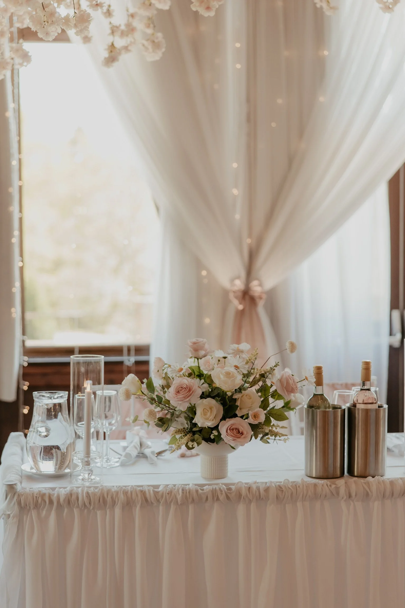 A wedding reception table with a pink and white floral arrangement, candles, and bottles of water or wine, set in front of sheer curtains and a window with trees outside.