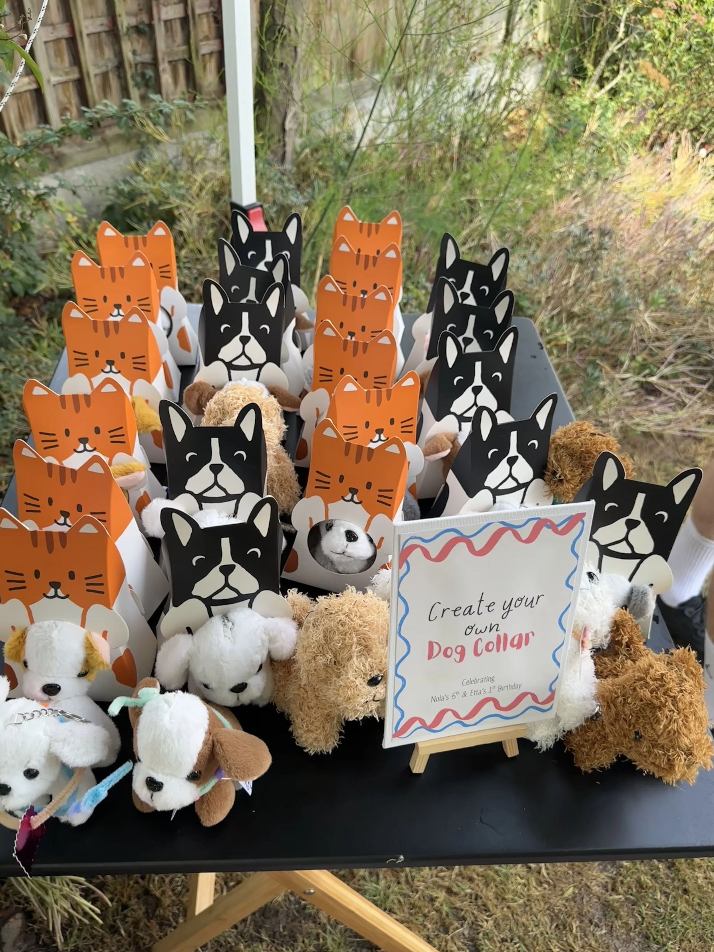 A table with plush dog toys and paper dog collars shaped like orange and black cats, featuring a sign that reads 'Create your own Dog Collar, Celebrating Nola's 5th & Elsa's 7th Birthday'.