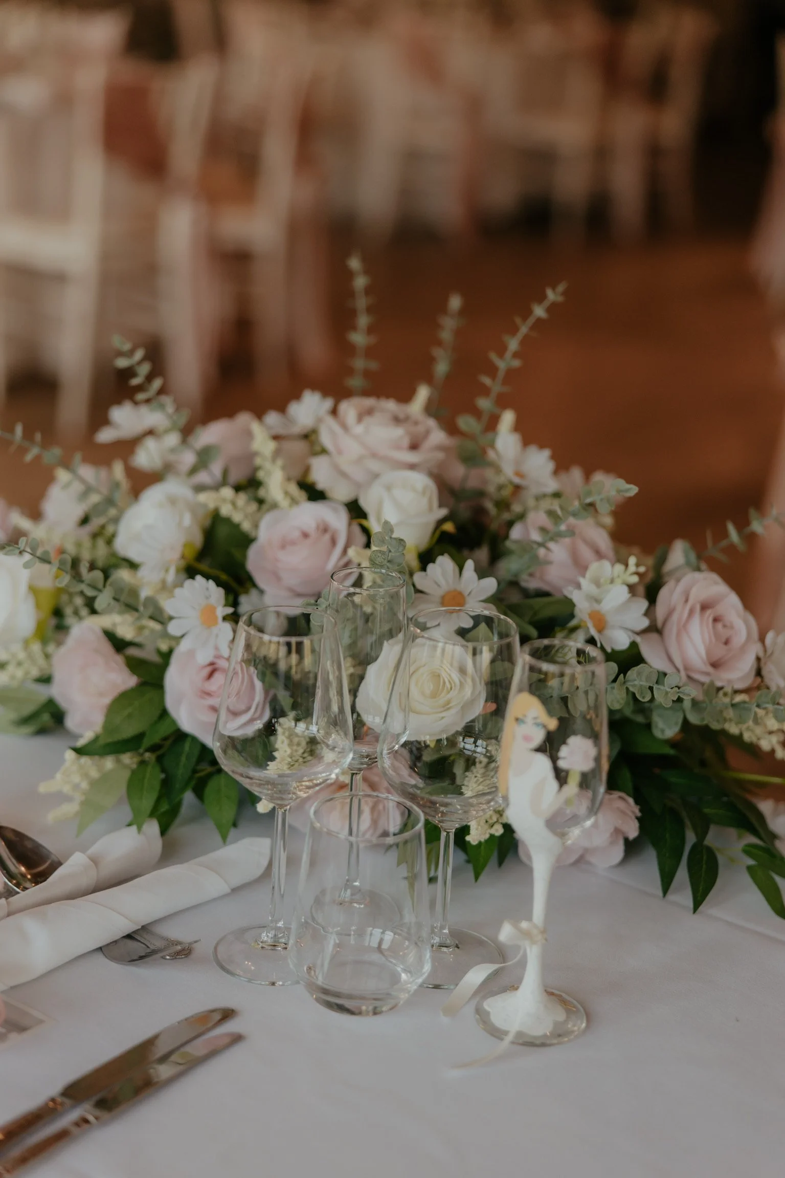 Elegant table setting with a large floral centerpiece of pale pink and white roses, daisies, and greenery. There are empty wine glasses, a water glass, and silverware on a white tablecloth, with a small figurine of a woman on a glass stand.