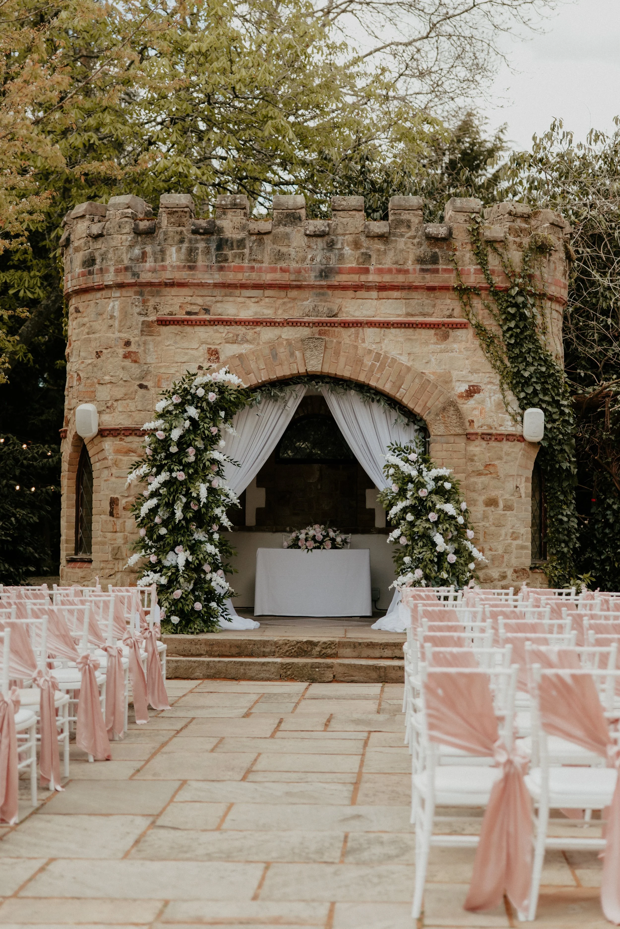 Outdoor wedding ceremony setup with rows of white chairs decorated with pink sashes, a floral arch with white and pink flowers, and a stone castle-like backdrop with greenery and white curtains.