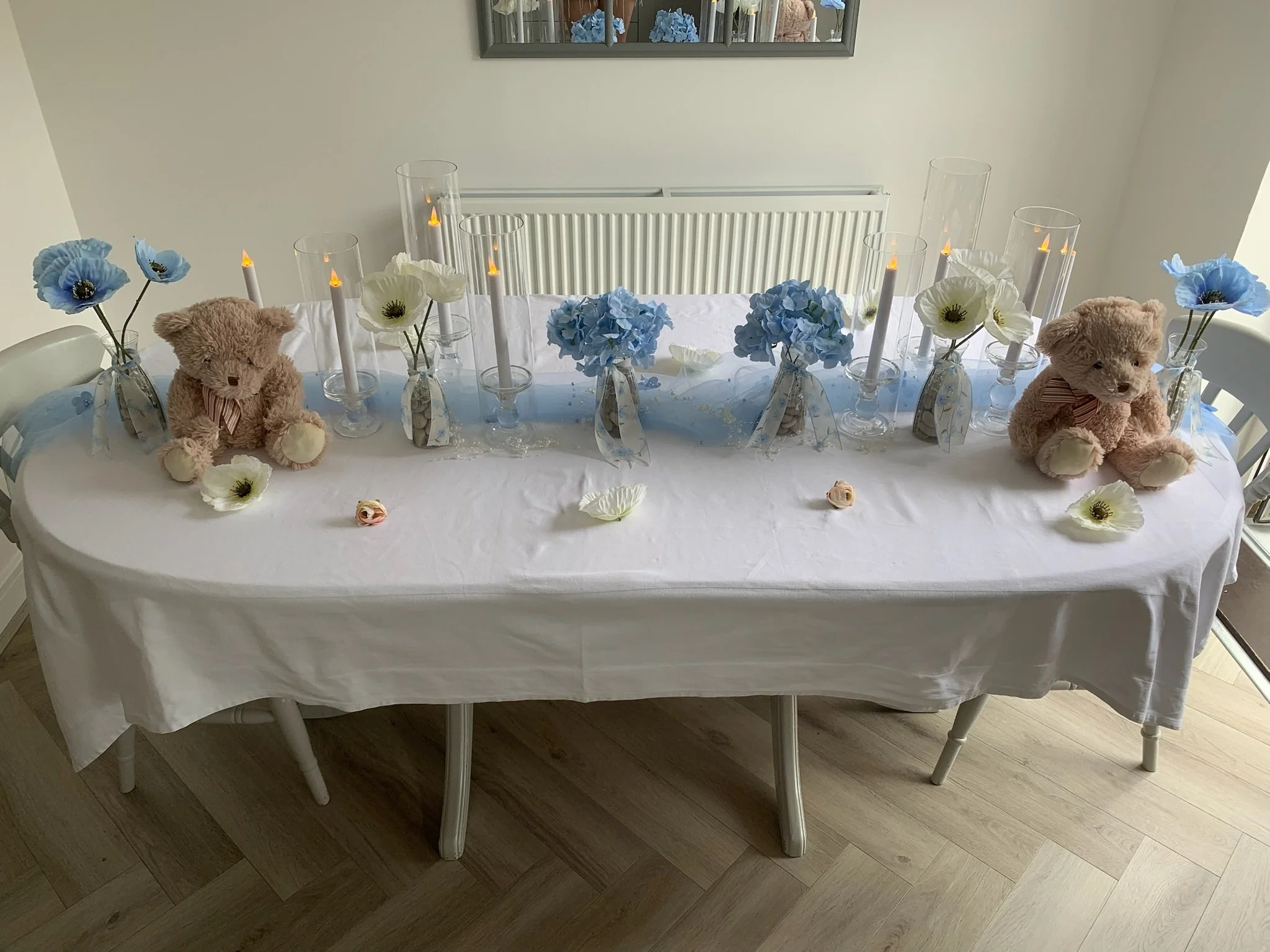 A dining table decorated with plush teddy bears, glass vases with blue and white artificial flowers, and candles in glass holders, set on a white tablecloth in a bright room.