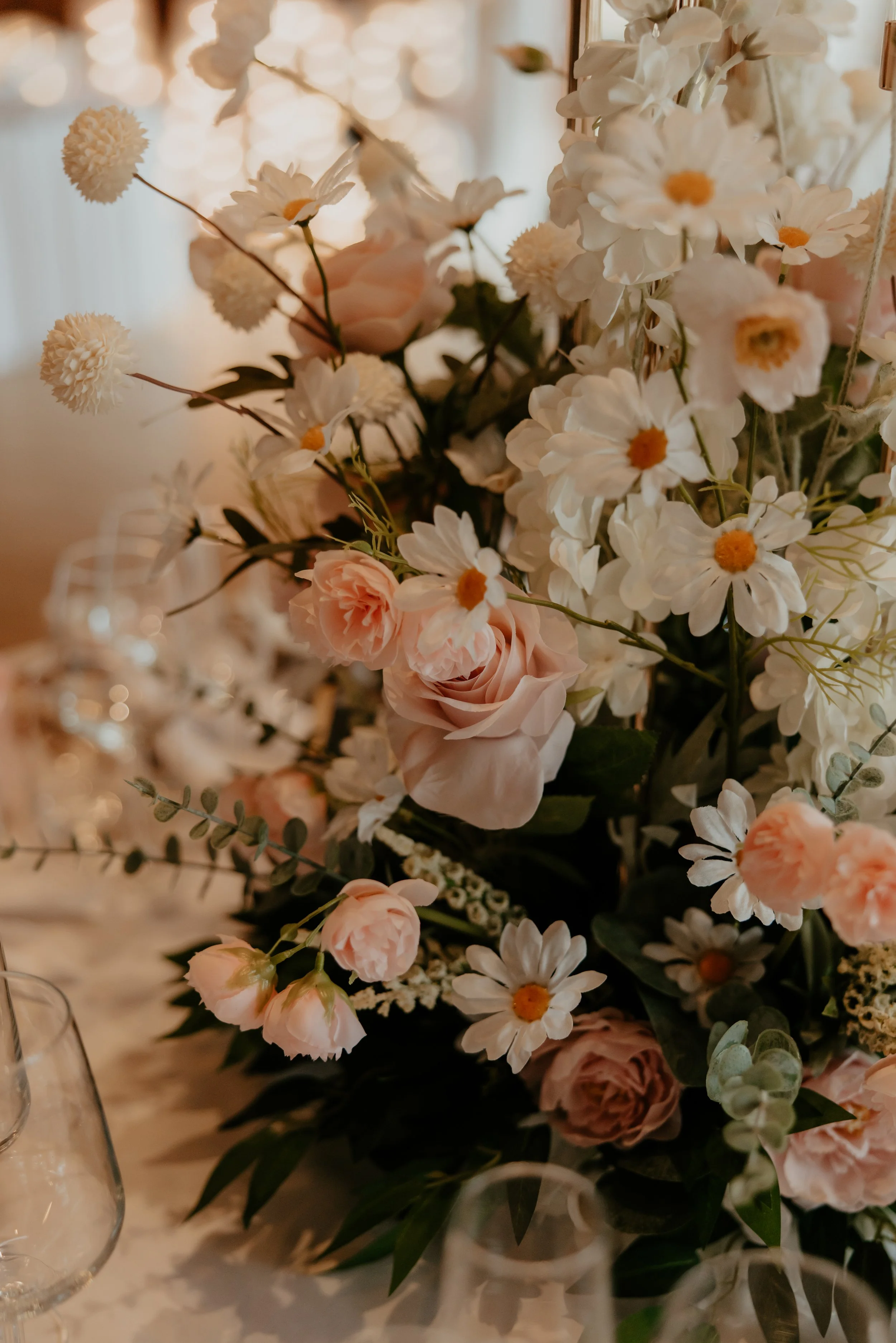 Close-up of a floral centerpiece with pink roses, white daisies, and greenery on a table, blurred background.