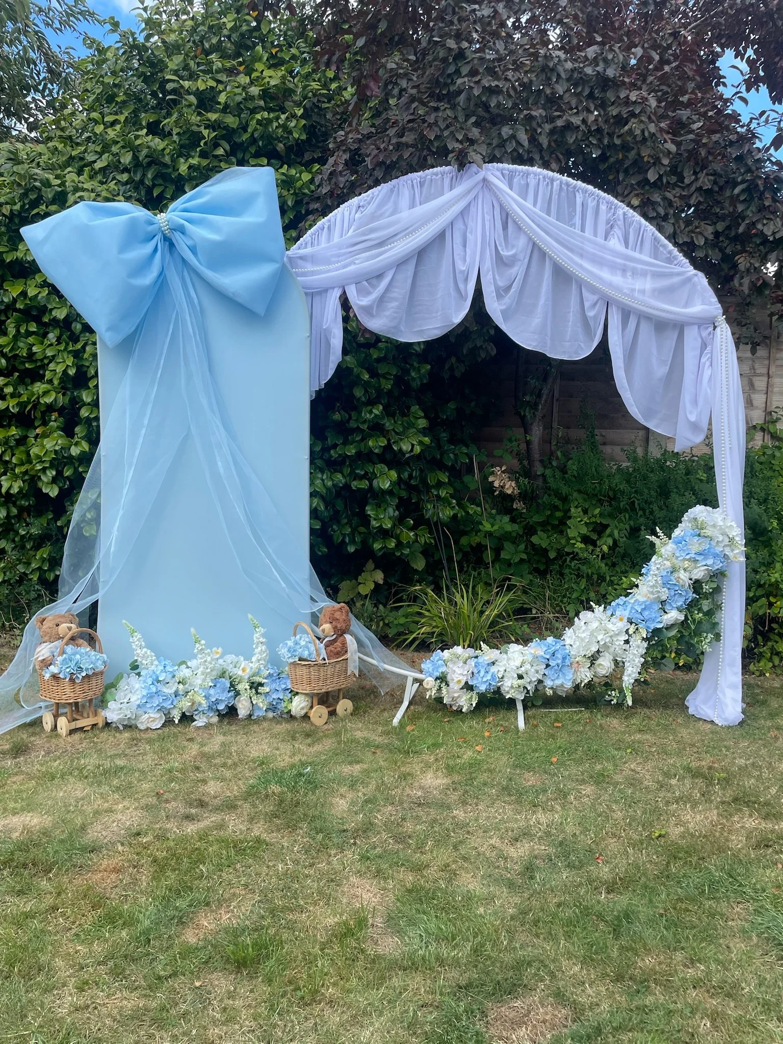An outdoor wedding arch decorated with white and blue flowers, draped white fabric, and a large blue bow, set against a background of greenery.