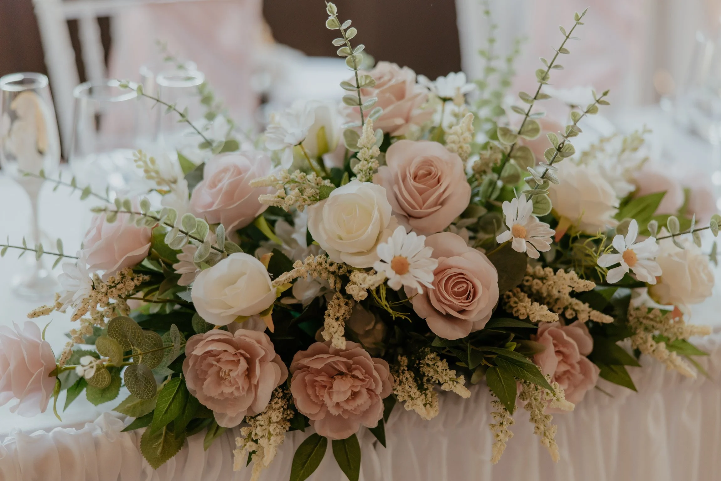 A floral centerpiece on a table with pink and white roses, daisies, and greenery. Champagne glasses are in the background.