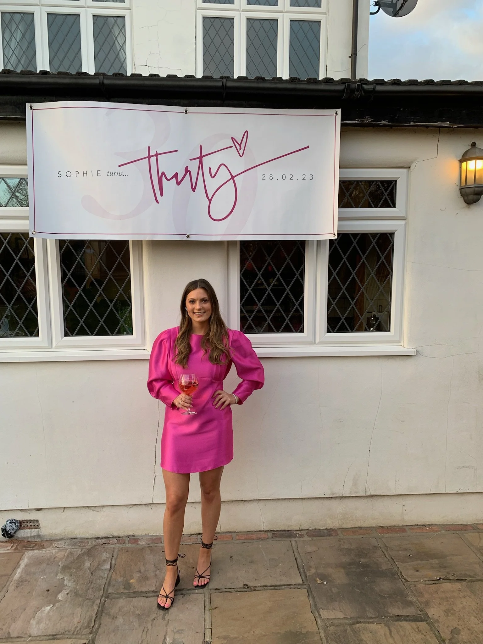 A young woman wearing a bright pink dress and black strappy heels is standing outside in front of a white wall with windows. She is smiling and holding a glass of pink drink. There is a large banner above her with the name 'Thirty' and information ab