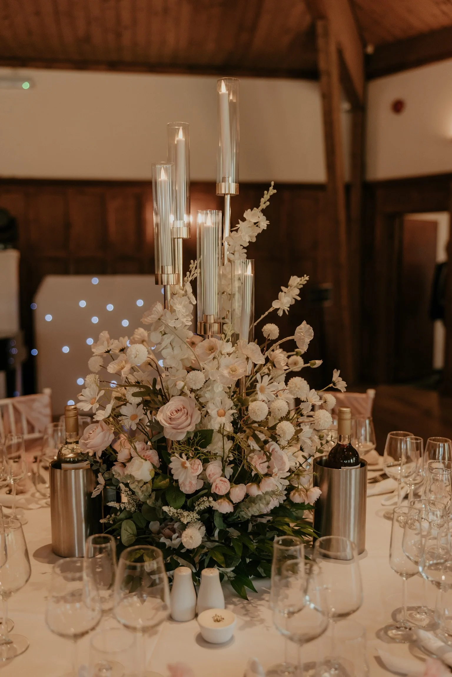 Elegant table centerpiece with white and blush roses, white daisies, and other flowers in a lush arrangement, accented with tall glass candle holders, candles, and bottles of wine, set on a formal dining table.