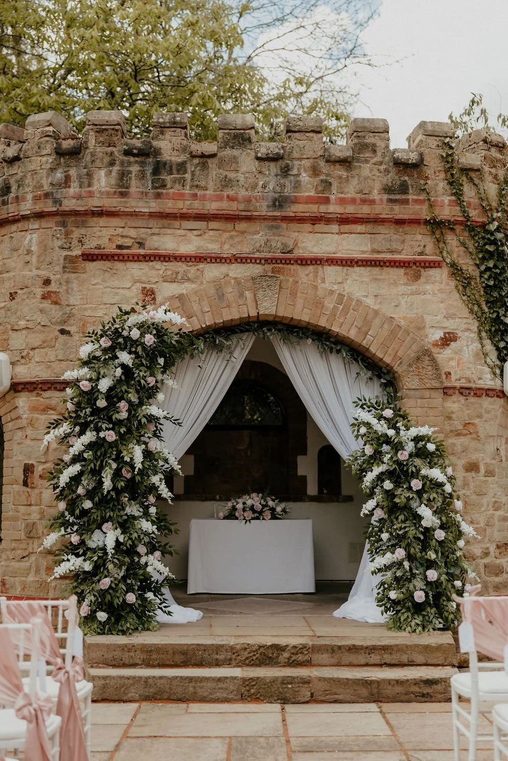 A rustic stone archway decorated with a lush floral arch of pink roses, white flowers, and greenery.