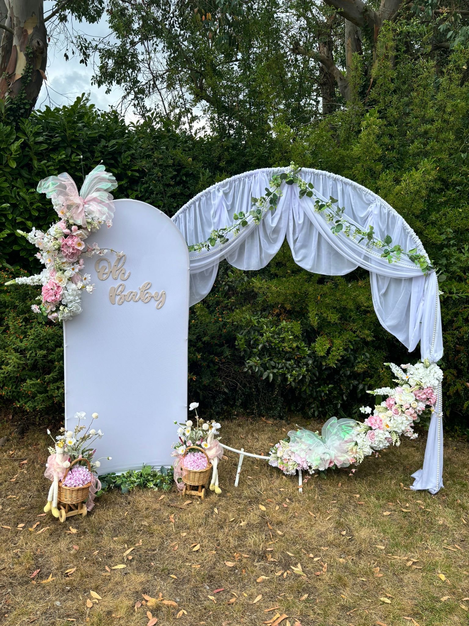 Baby shower decoration with a white floral arch and a tall white sign that says 'Oh Baby'. The sign is decorated with pink and white flowers and pastel-colored butterfly embellishments. Small wicker baskets filled with pink and white flowers are plac
