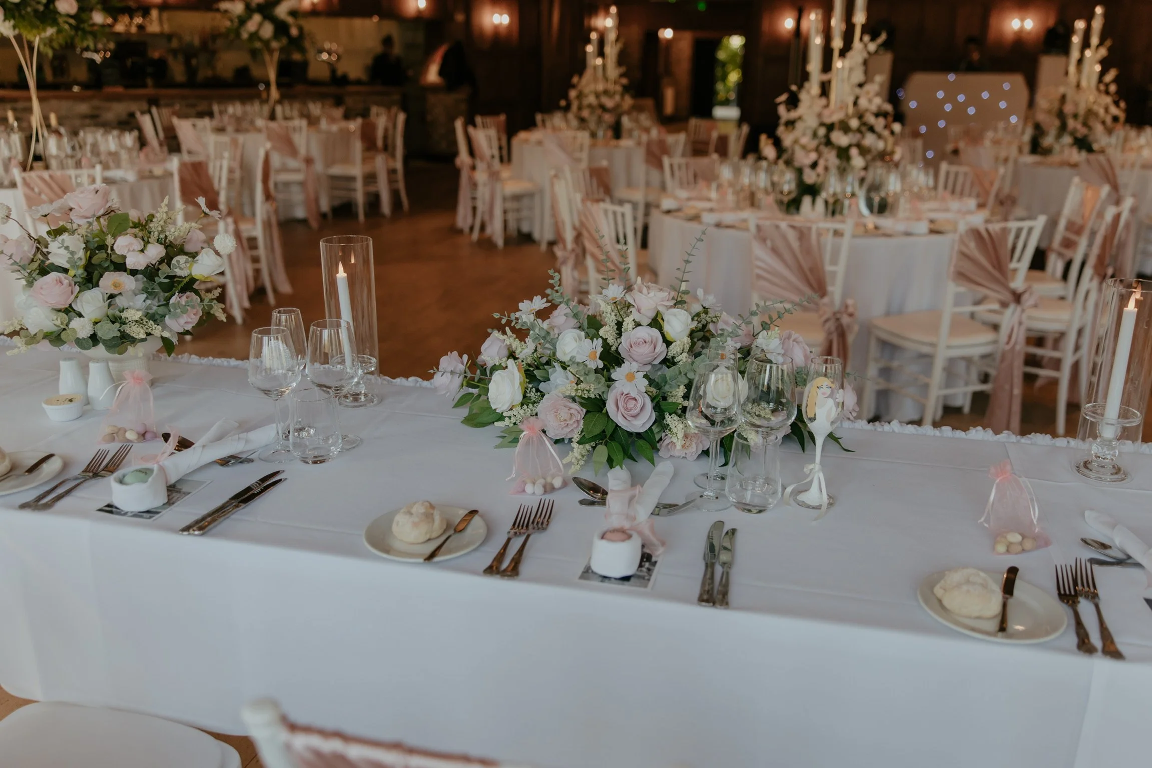 A shot from behind the top table. Centre piece floral arrangement with white roses and blush pink flowers on a wedding top table.