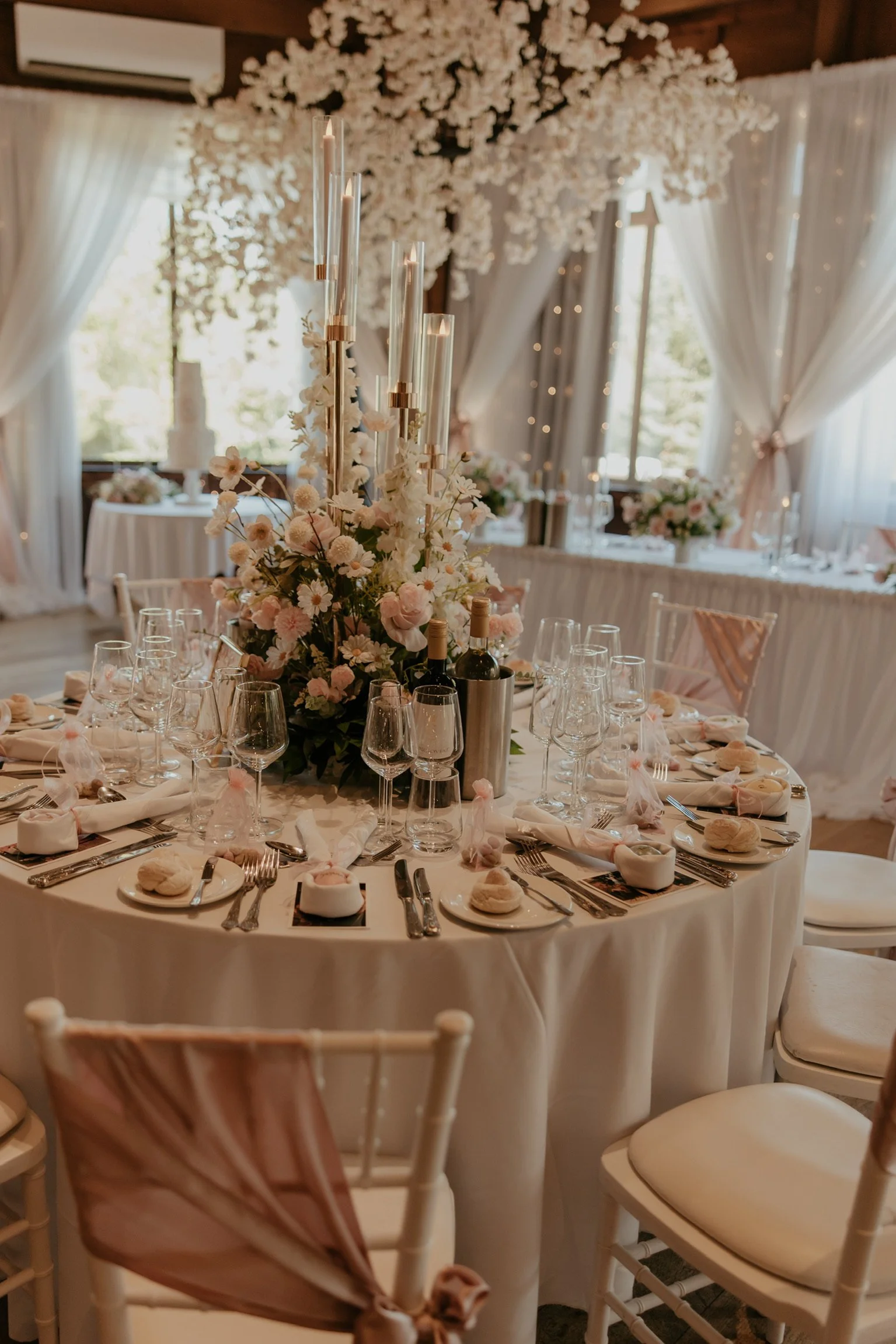 Elegant banquet table decorated with a large floral centerpiece, candles, wine bottles, and place settings in a decorated reception hall with draped curtains and windows.