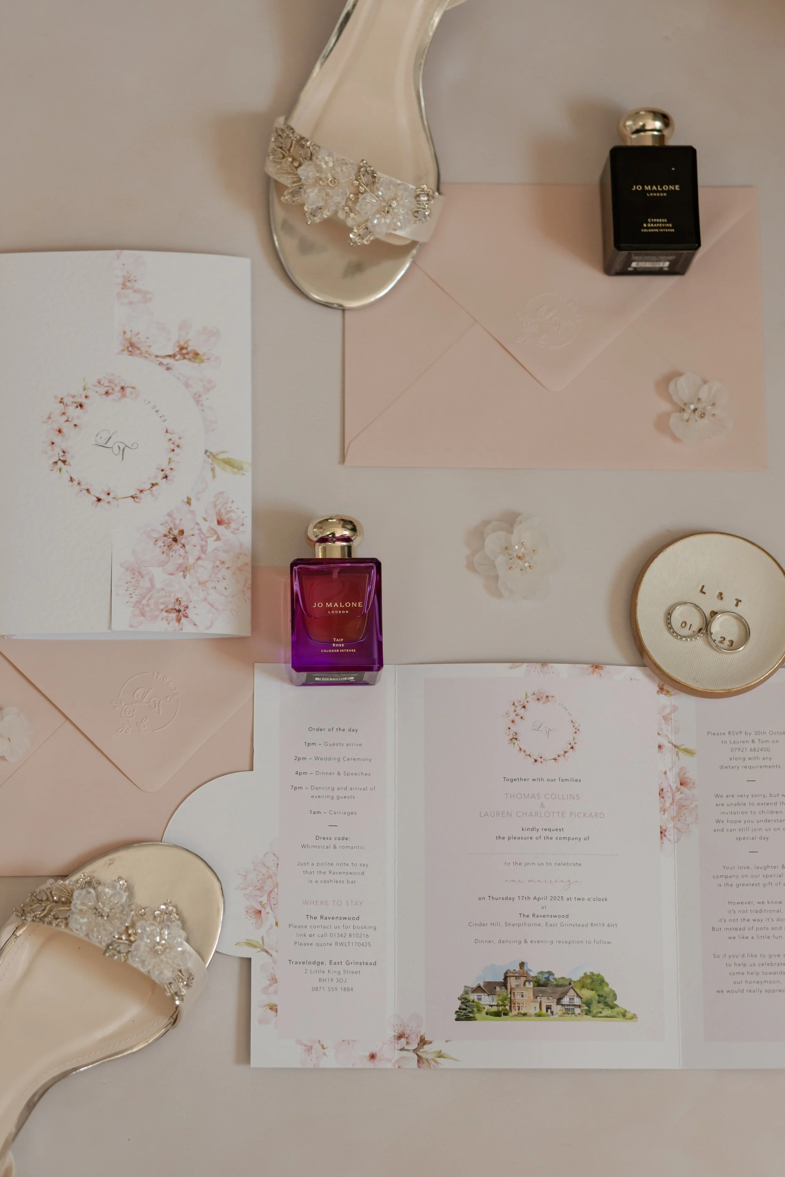 Wedding invitation, two bottles of Jo Malone perfume, ivory bridal shoes with floral embellishments, pink envelope with embossed heart, white flower decorations, and a wedding menu on a pink and white table.