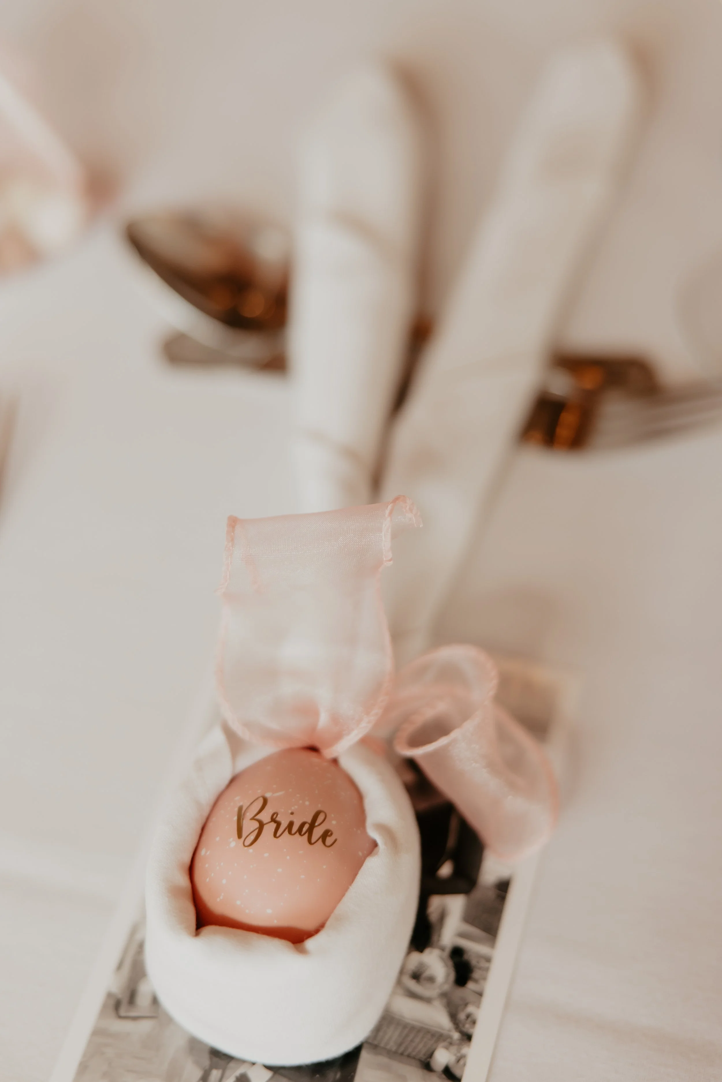 A pale pink decorative egg with the word 'Bride' in gold lettering, wrapped in a light pink satin ribbon, resting in a white ring holder.