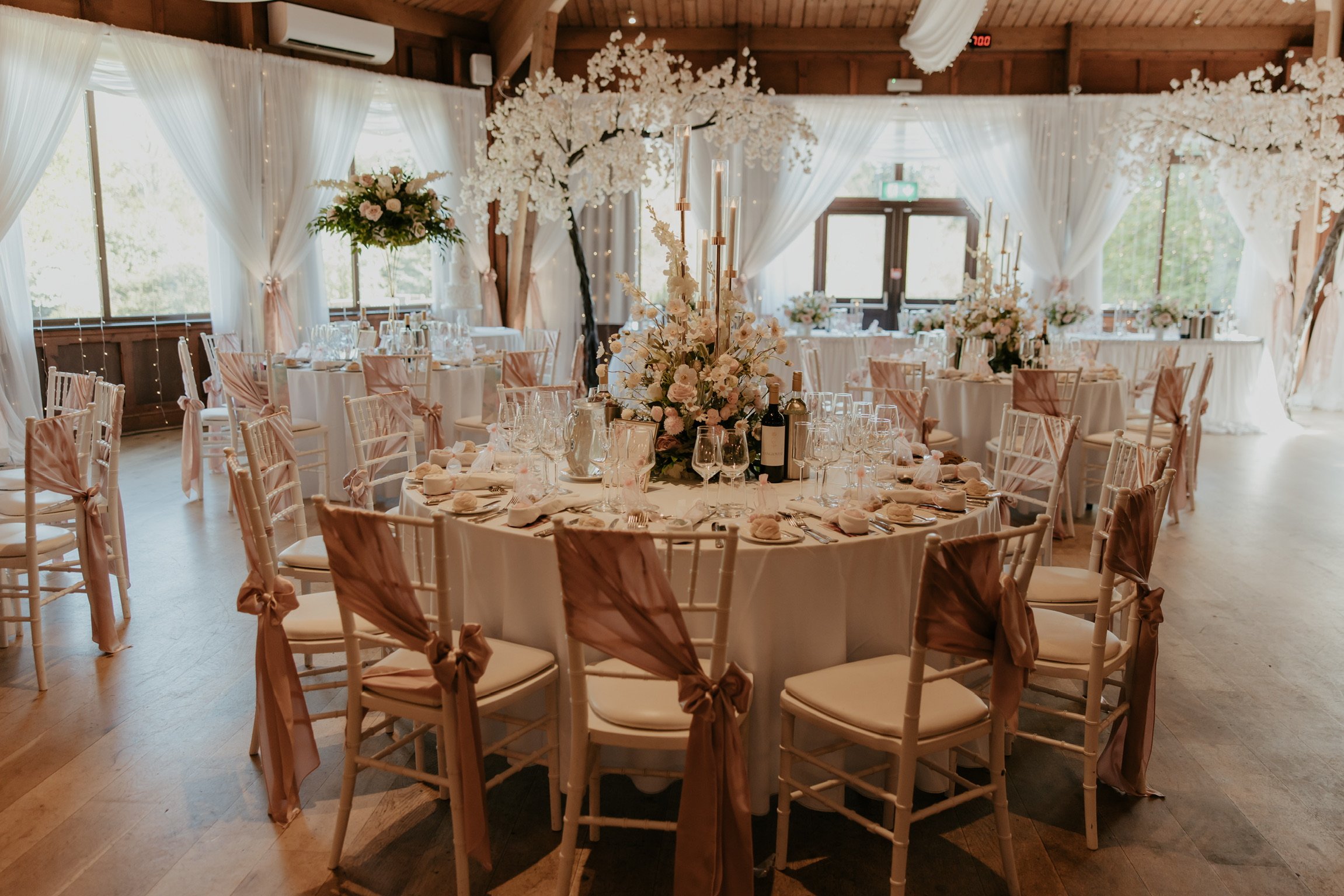 Elegant banquet hall decorated for a wedding reception with round tables, white tablecloths, and tall floral centerpieces. Chairs are adorned with pink ribbons. The room has large windows, white curtains, and decorative trees with white blossoms.
