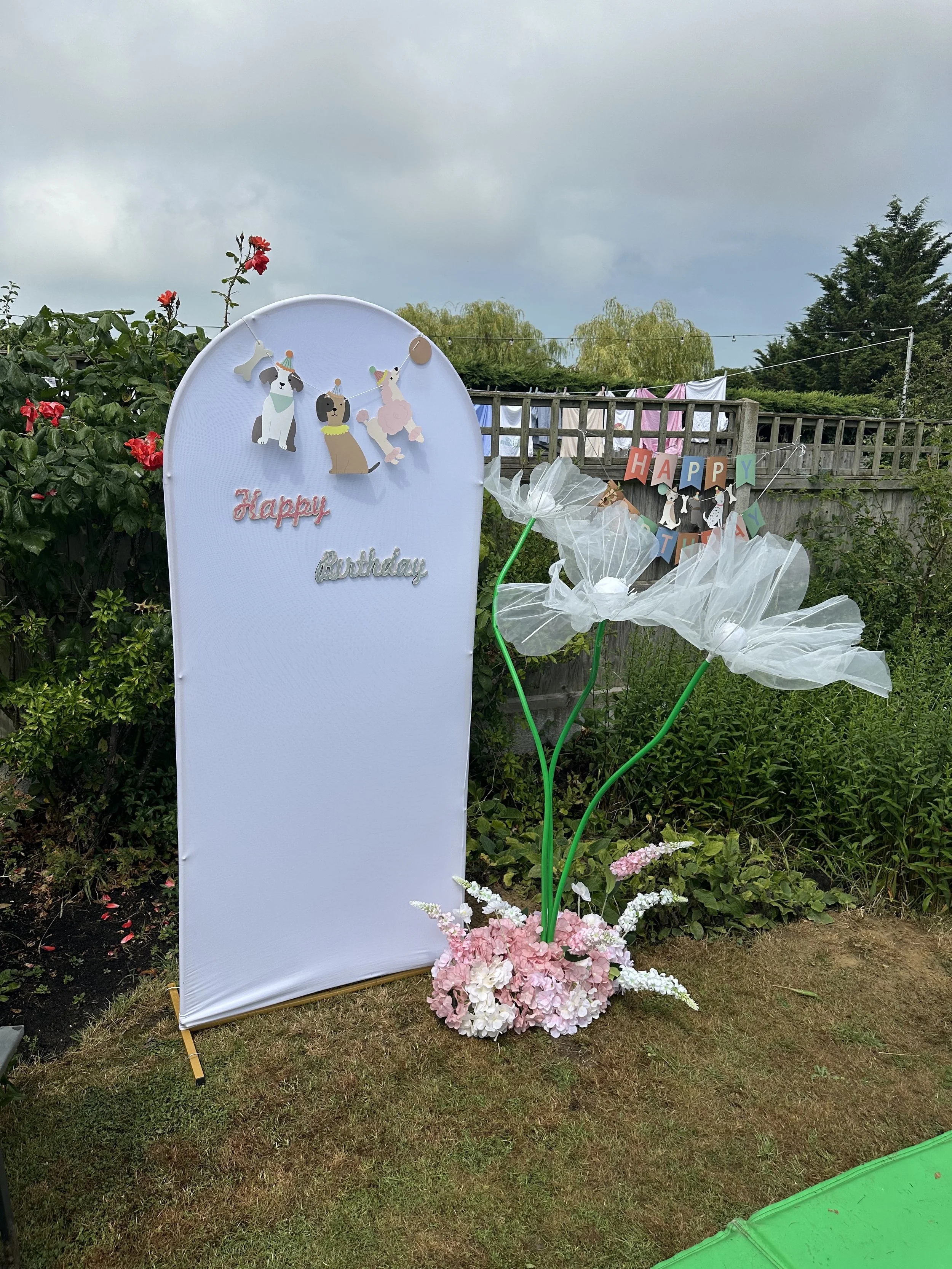 Outdoor birthday decoration with a white sign that says 'Happy birthday' and a pink floral arrangement at the base. Next to the sign, there are large white tissue paper flowers on green stems with a 'Happy Birthday' banner in the background. The scen