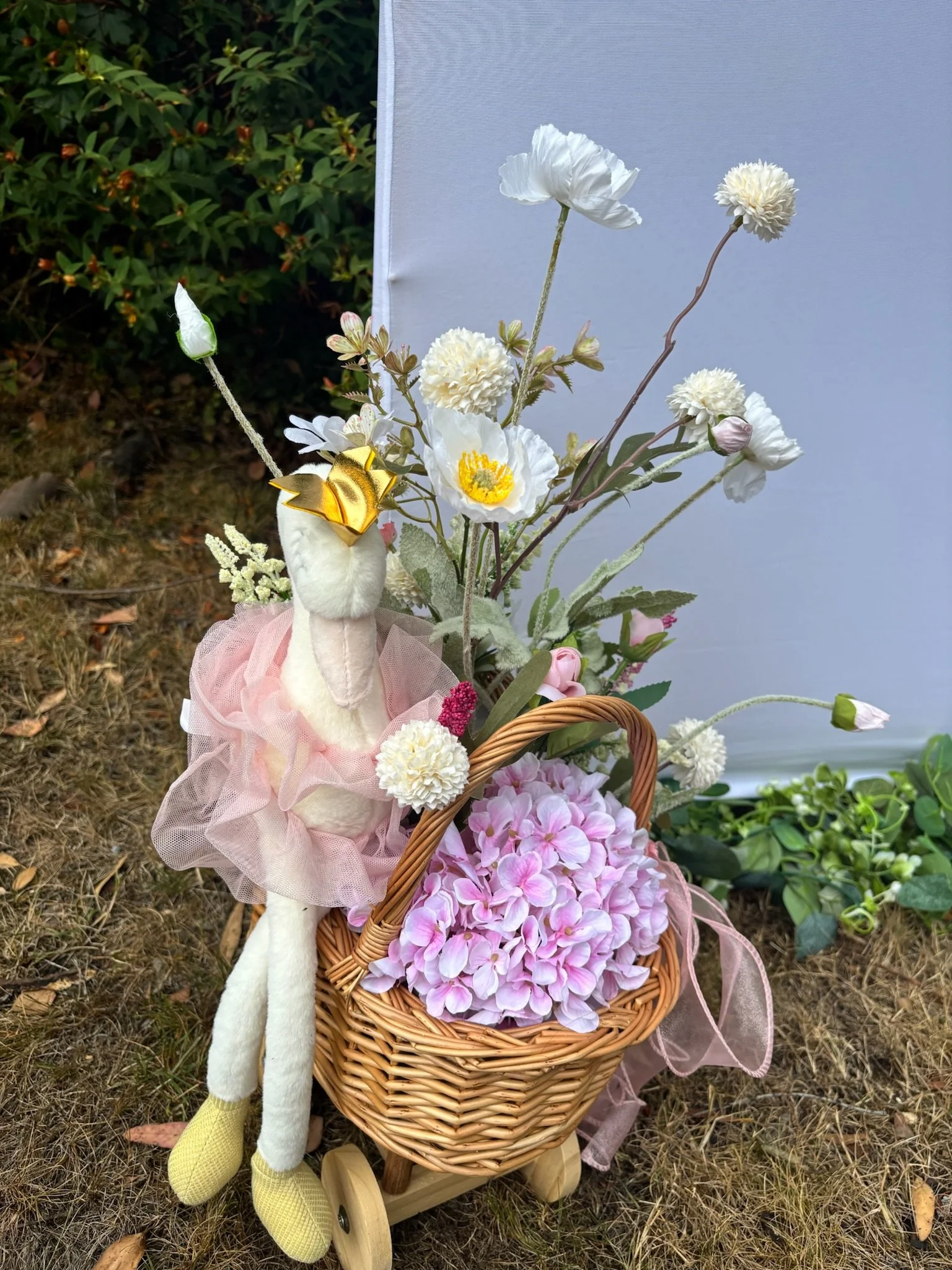 A decorative arrangement of white and pink flowers in a wicker basket, accompanied by a plush toy swan with a pink tutu and yellow shoes, placed outdoors on a grassy area with green foliage in the background.