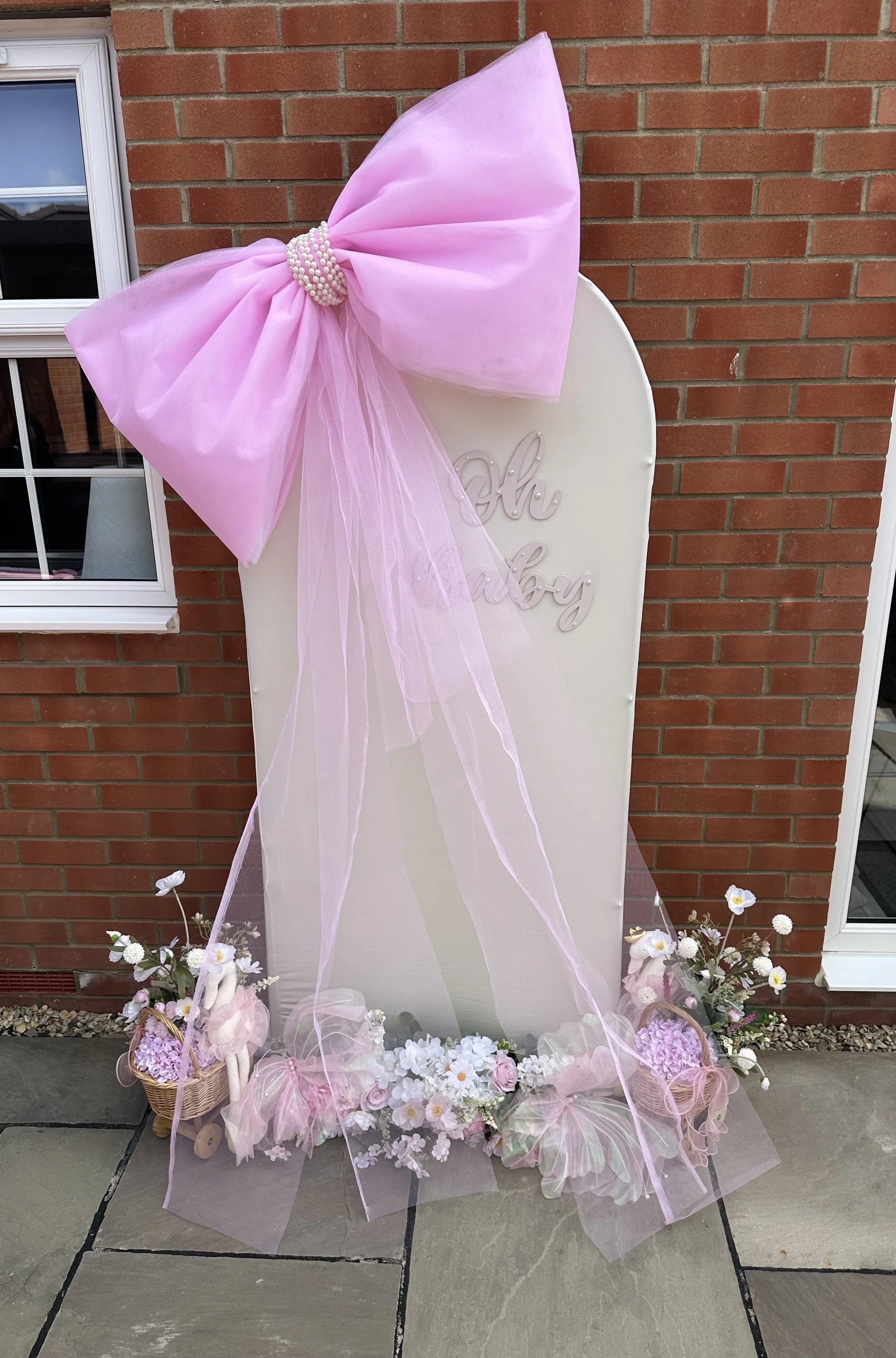 A decorative standing board with the words 'Oh Baby' and adorned with a large pink bow, netting, and surrounded by pink and white flowers on a patio in front of a brick wall.