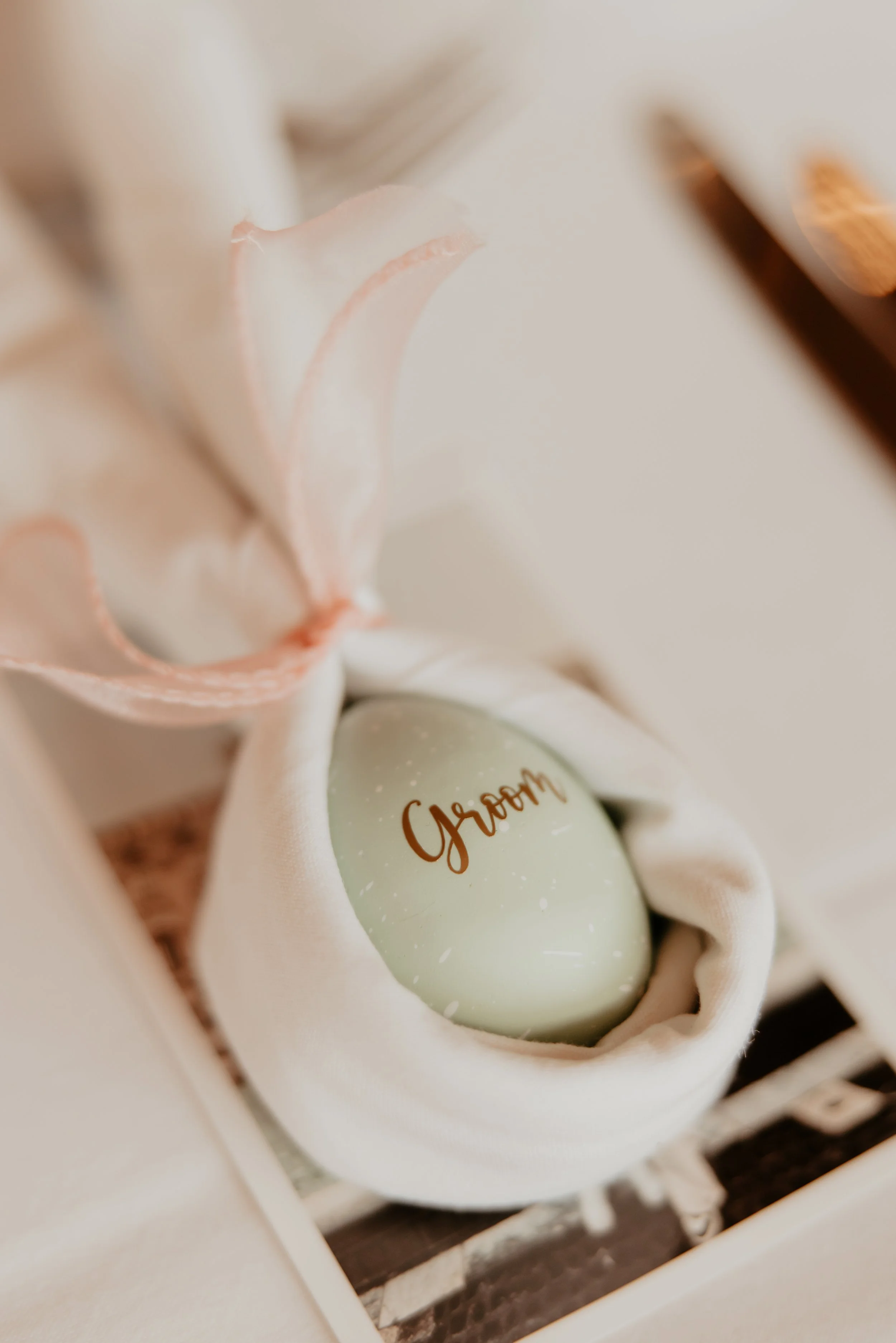A light green decorative egg with the word "Groom" written on it in brown, nestled in white fabric with a pink ribbon, inside a white box with shredded paper.