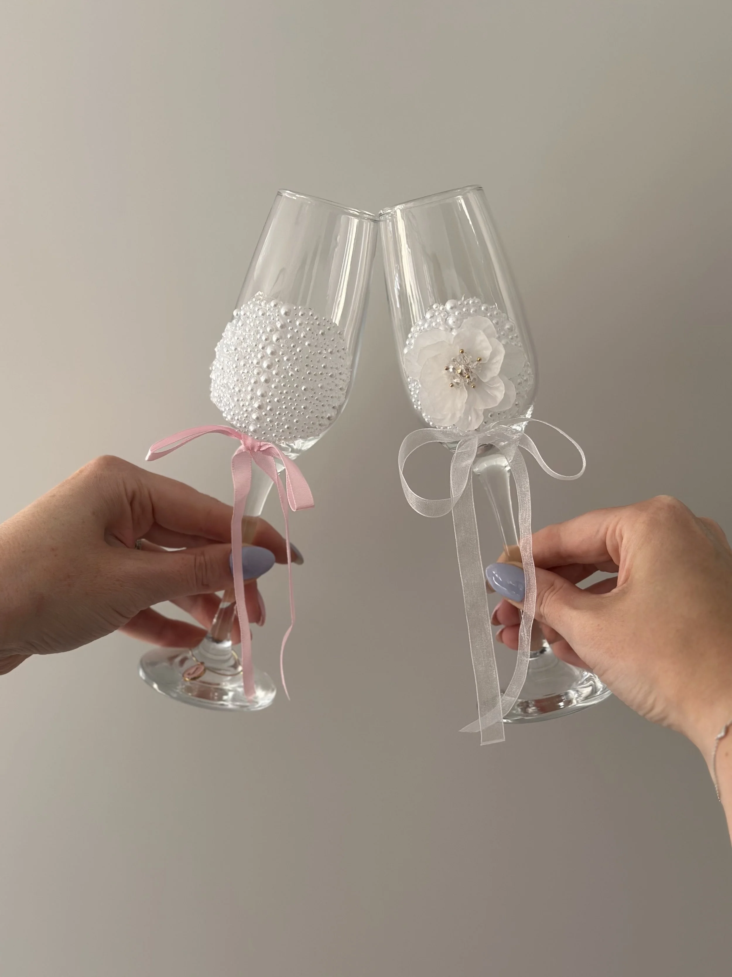 Two hands holding champagne flutes decorated with white ribbons and fabric flowers, with one flute decorated with pearls and the other with a flower and rhinestones.
