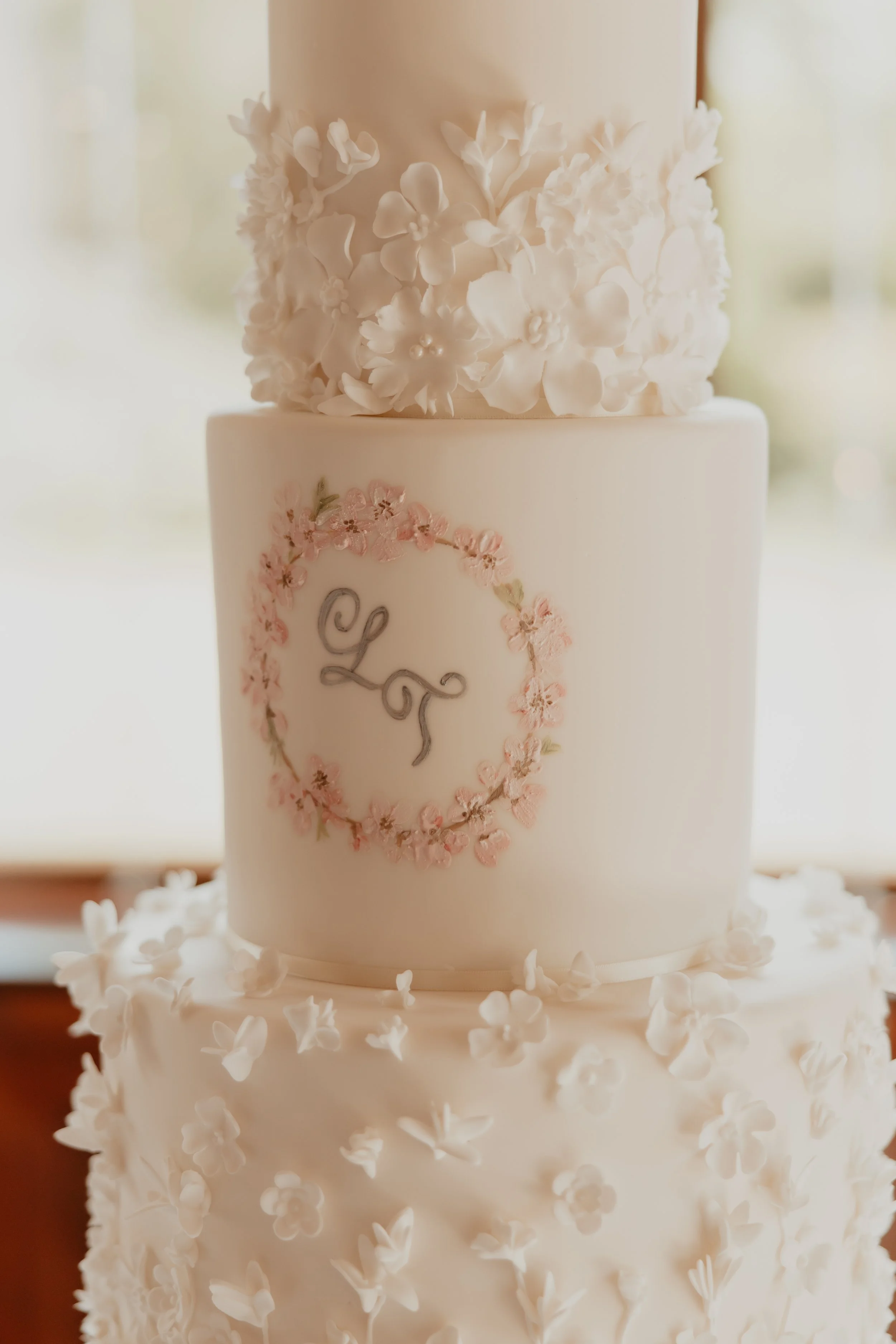 A three-tier white wedding cake decorated with white floral accents on the top and bottom tiers, and a pink floral monogram in the center of the middle tier.