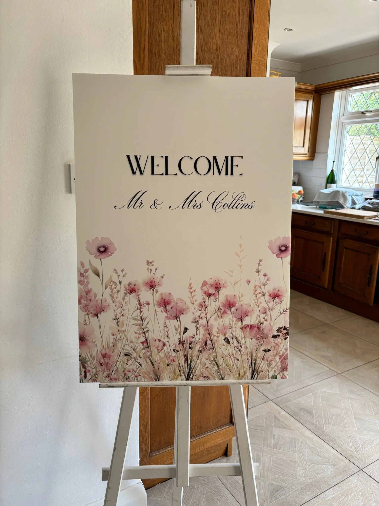 Welcome sign with floral design reading 'Welcome Mr & Mrs Collins' on an easel in a kitchen.