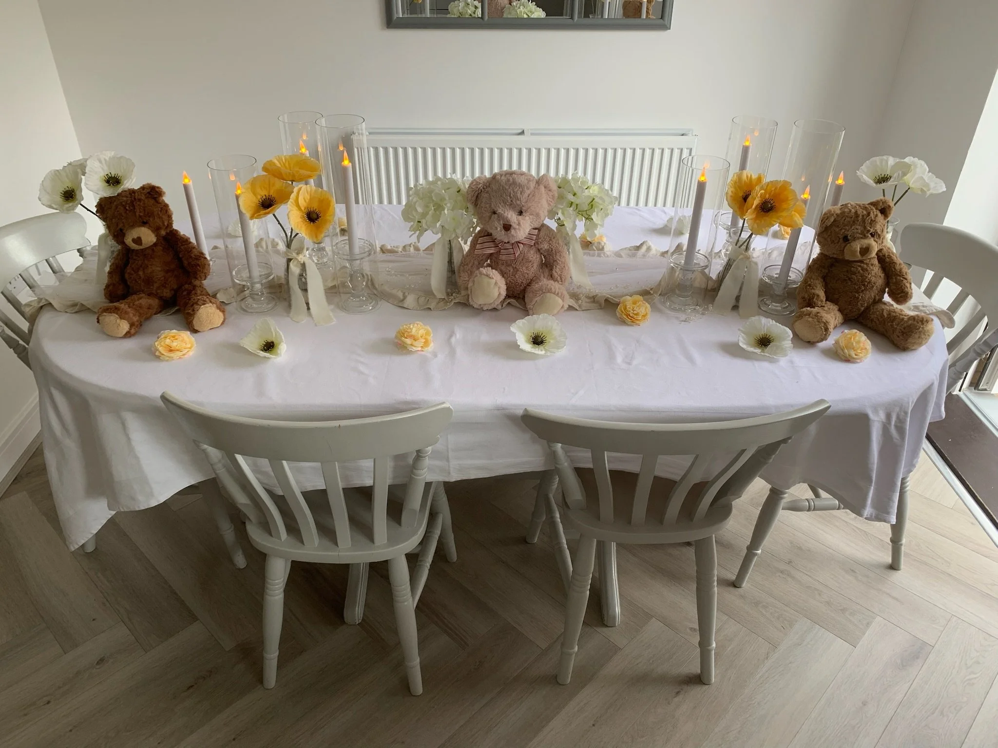 A decorated dining table with a white tablecloth, four teddy bears, floral arrangements in white and yellow, and lit candles in glass holders.
