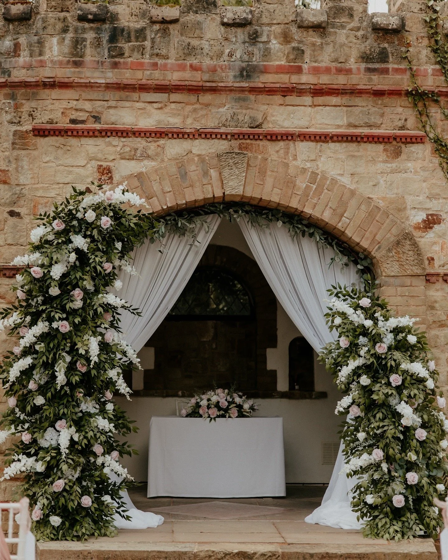 Wedding arch decorated with white and light pink flowers and green foliage, set against a brick wall with white curtains.