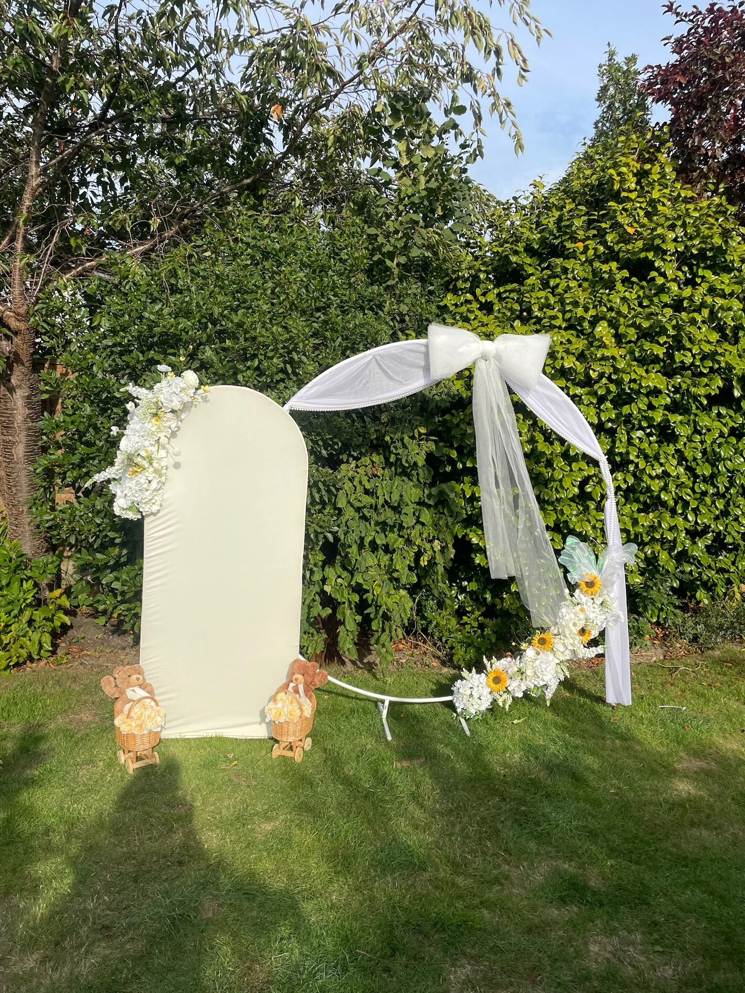 A white decorative wedding backdrop with floral arrangements and teddy bears on a grassy outdoor setting.