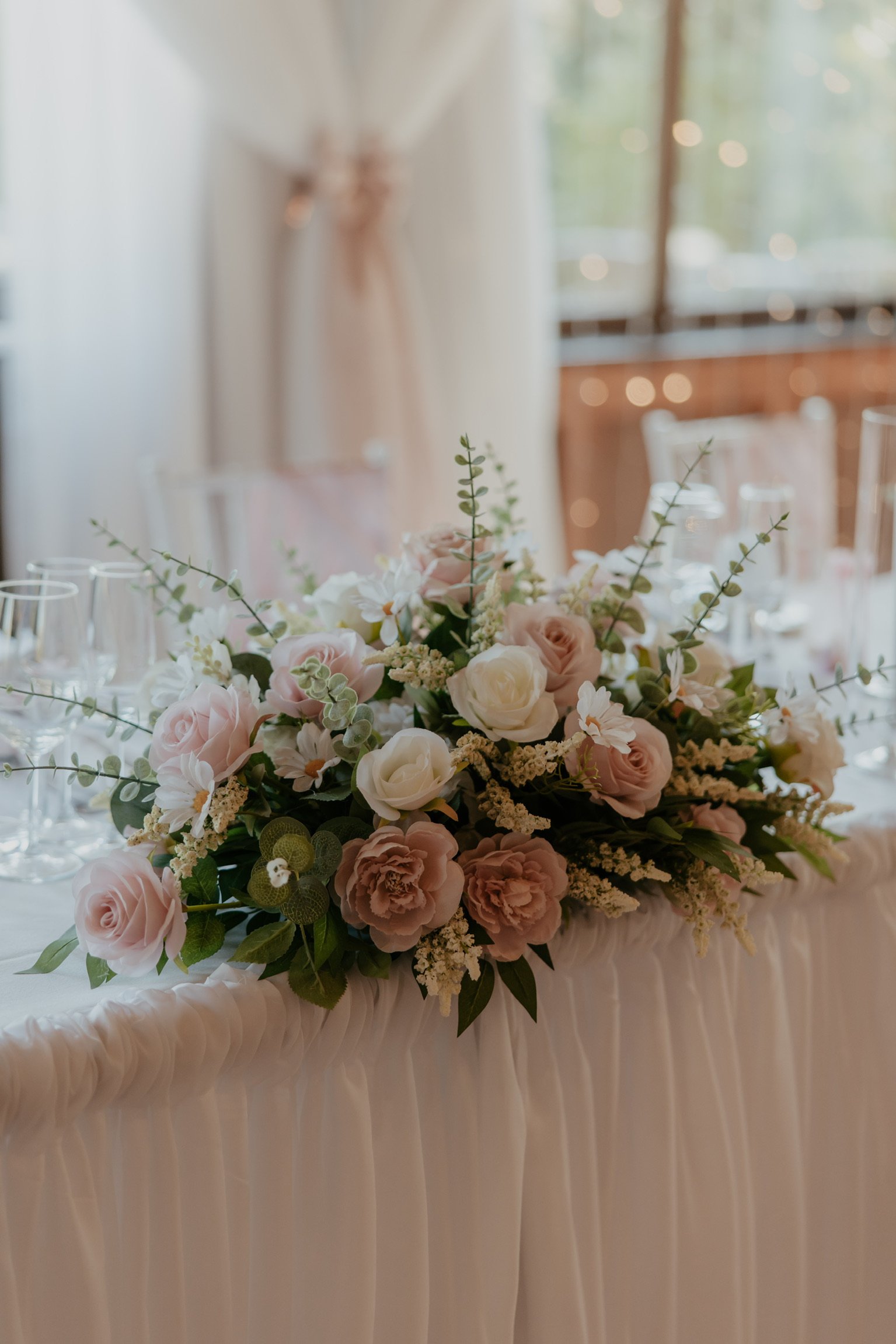 A floral centerpiece with pink and white roses, white daisies, and greenery on a table with a white ruffled tablecloth, set in a decorated event space with blurred background lighting and windows.