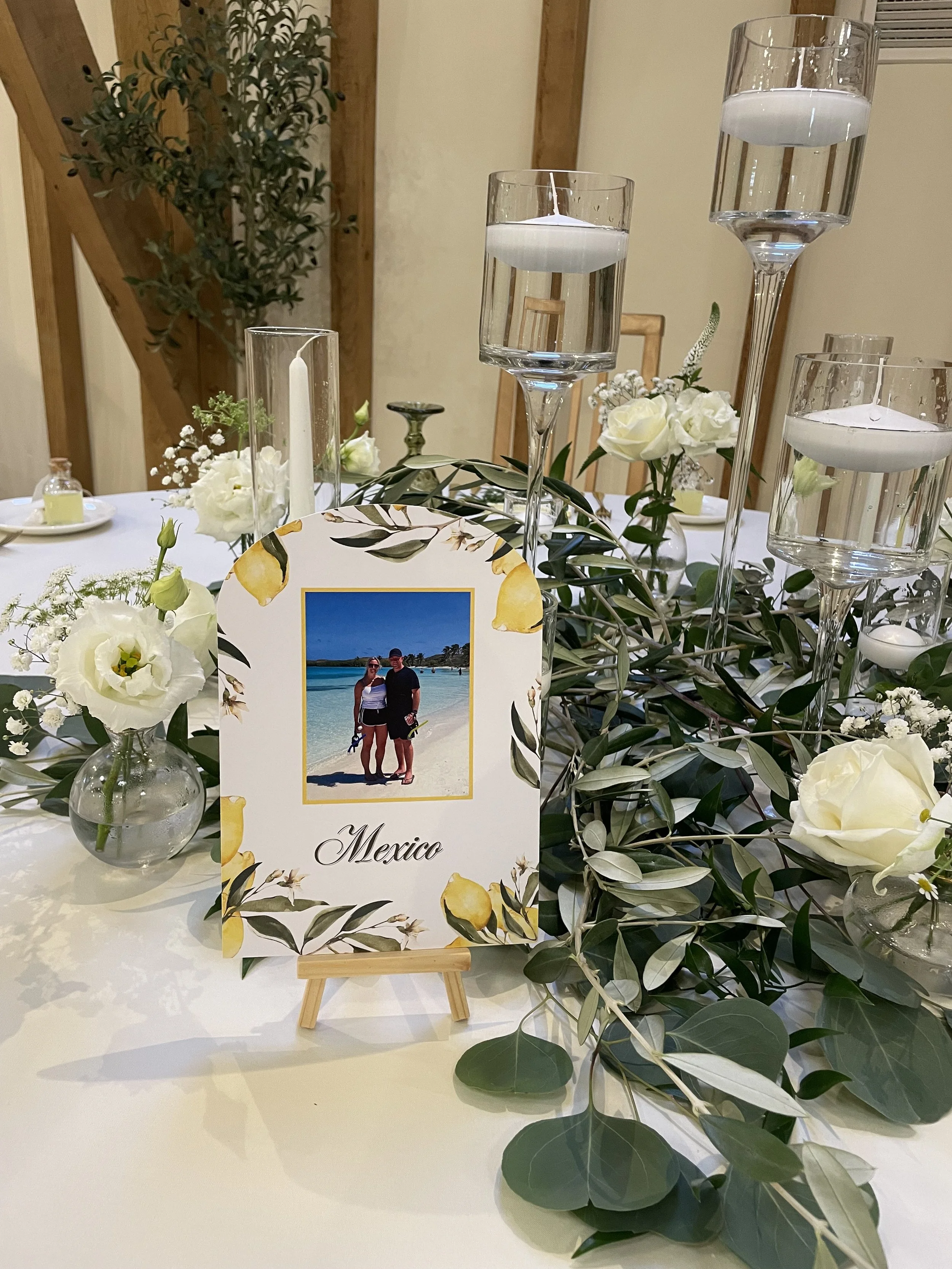 Table centerpiece with white flowers, greenery, candles in glass holders, and a photo card labeled Mexico by a small easel on a white tablecloth.
