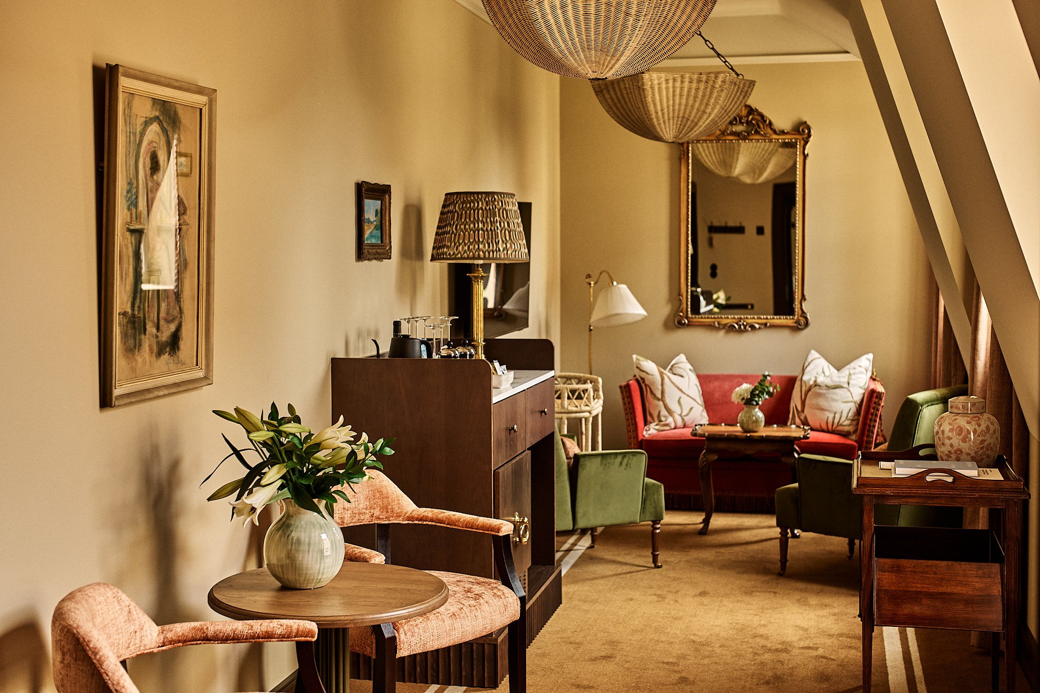 Interior of a living room with vintage-style furniture, wall art, and decorative lighting fixtures, featuring a sitting area with a red sofa, armchairs, and a coffee table.