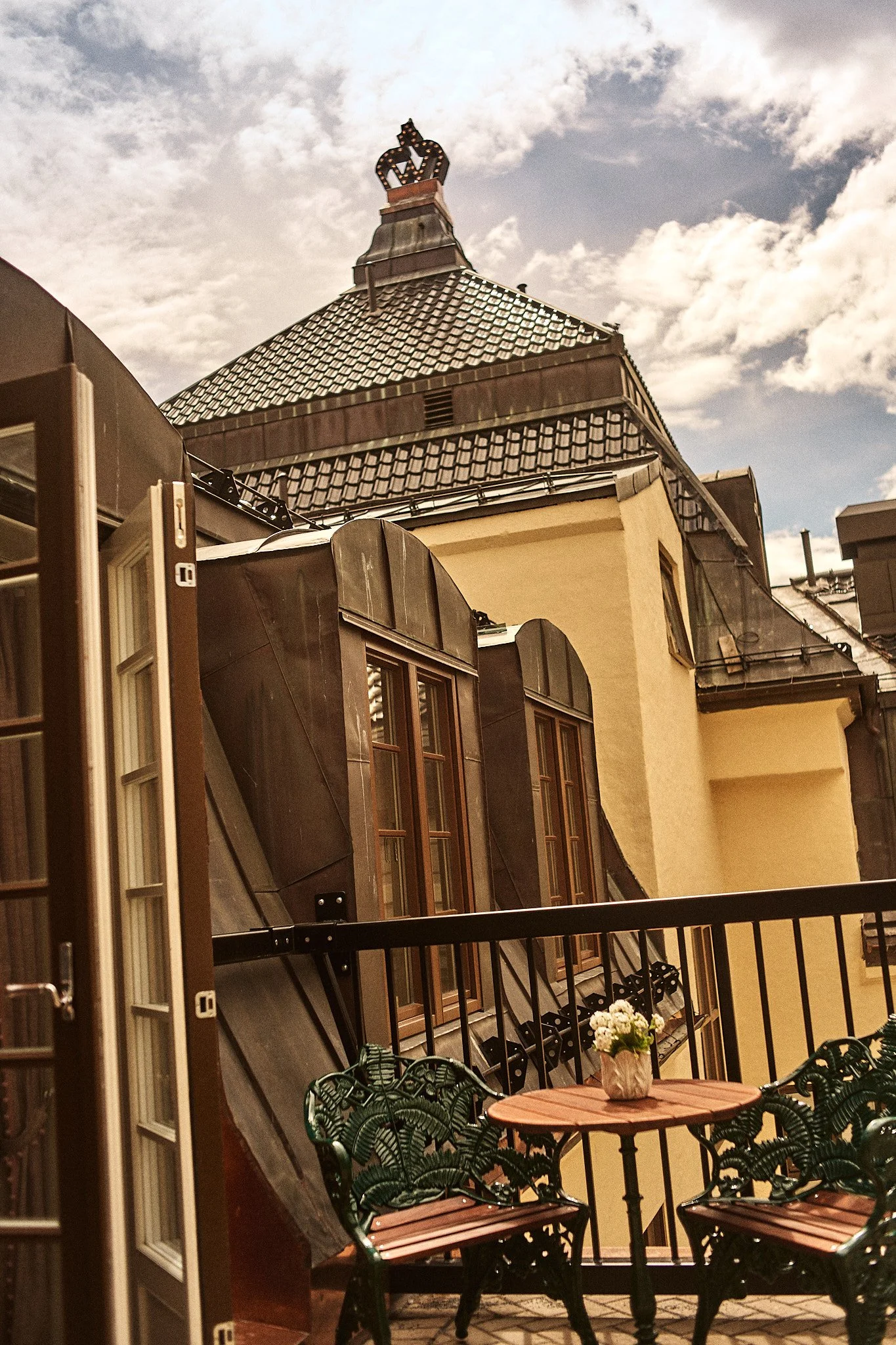 A balcony scene with an outdoor table and chairs, a potted plant with white flowers, view of older buildings with mansard roofs, and a cloudy sky.