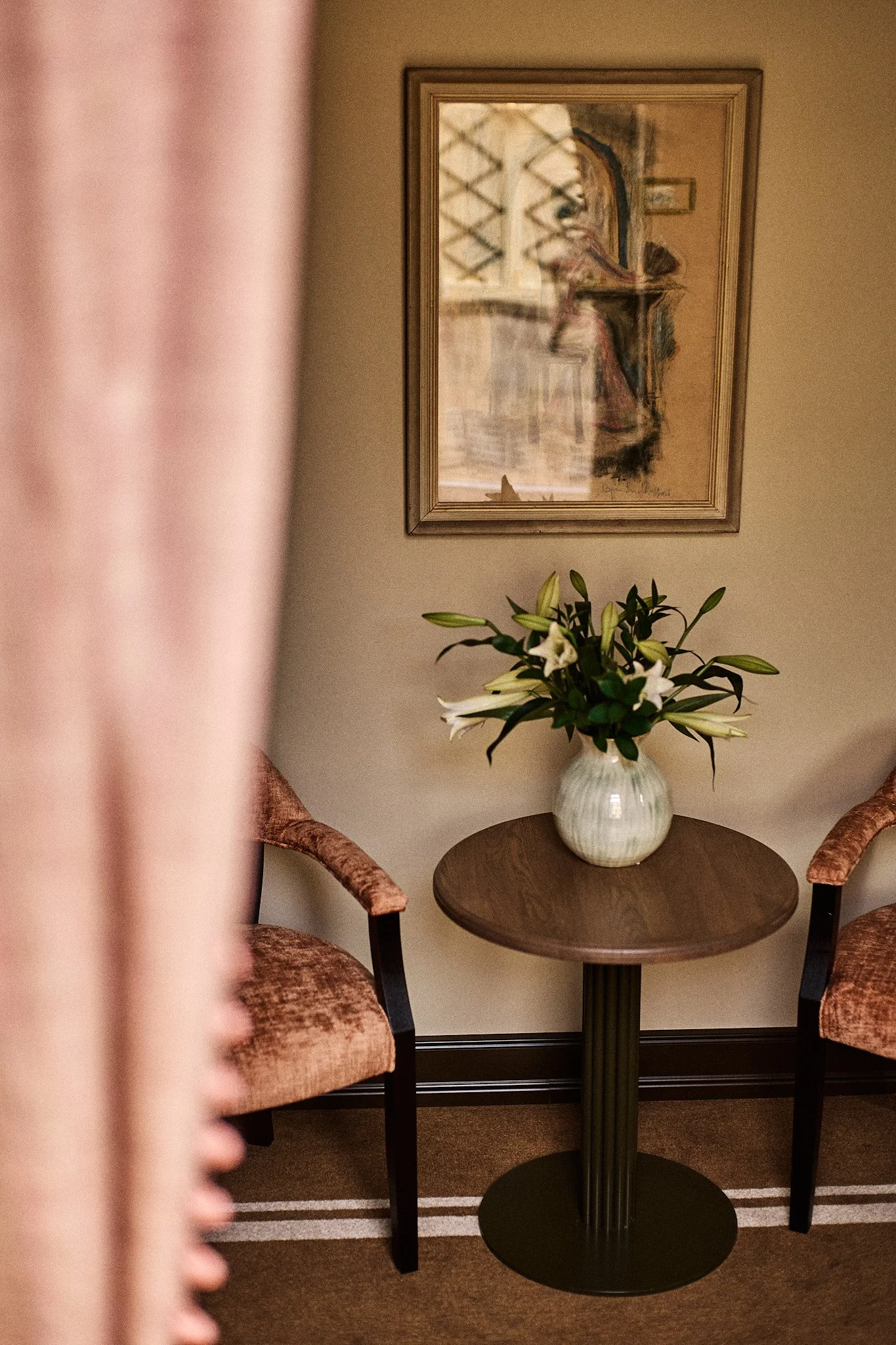 A small round wooden table with a black ribbed column pedestal base, holding a white vase with white lilies and greenery. Two upholstered armchairs with pinkish floral fabric are partially visible on either side of the table. On the wall behind, there is framed abstract art. The photograph is taken indoors, with pink curtains partially obscuring the left side of the image.