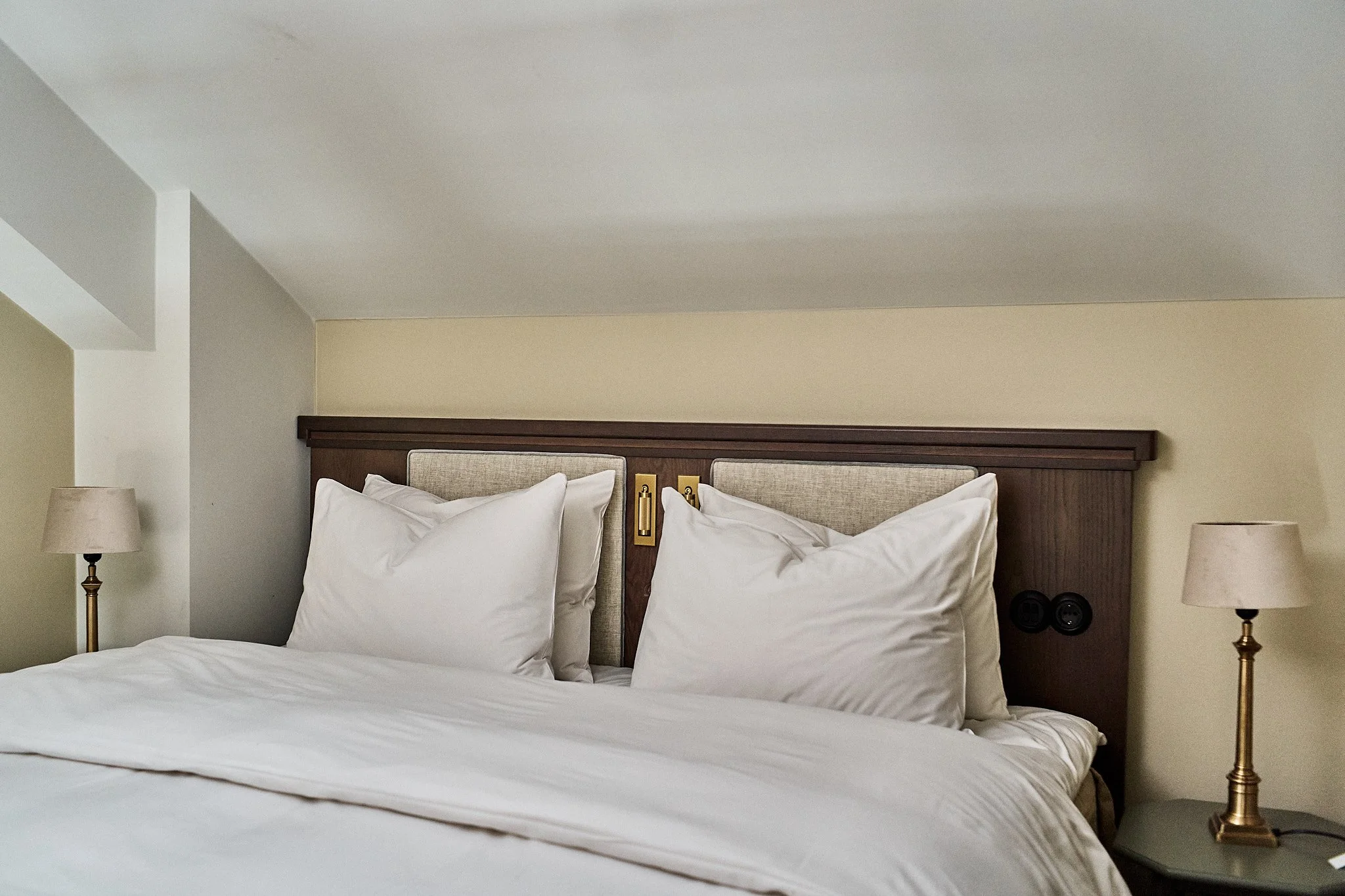 A neatly made hotel bed with white sheets and multiple pillows, a wooden headboard with beige fabric accents, two brass bedside lamps, and wall outlets on either side.