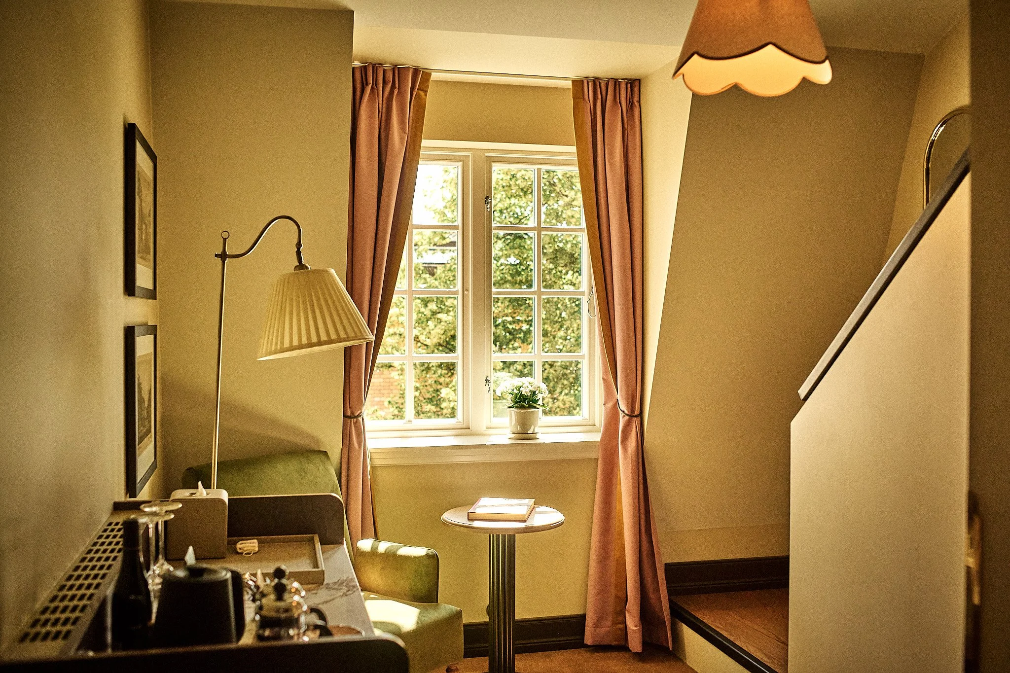A cozy room corner with a window framed by pink curtains, a small table with a book, a green upholstered chair, a floor lamp with a pleated shade, and a hanging ceiling lampshade.