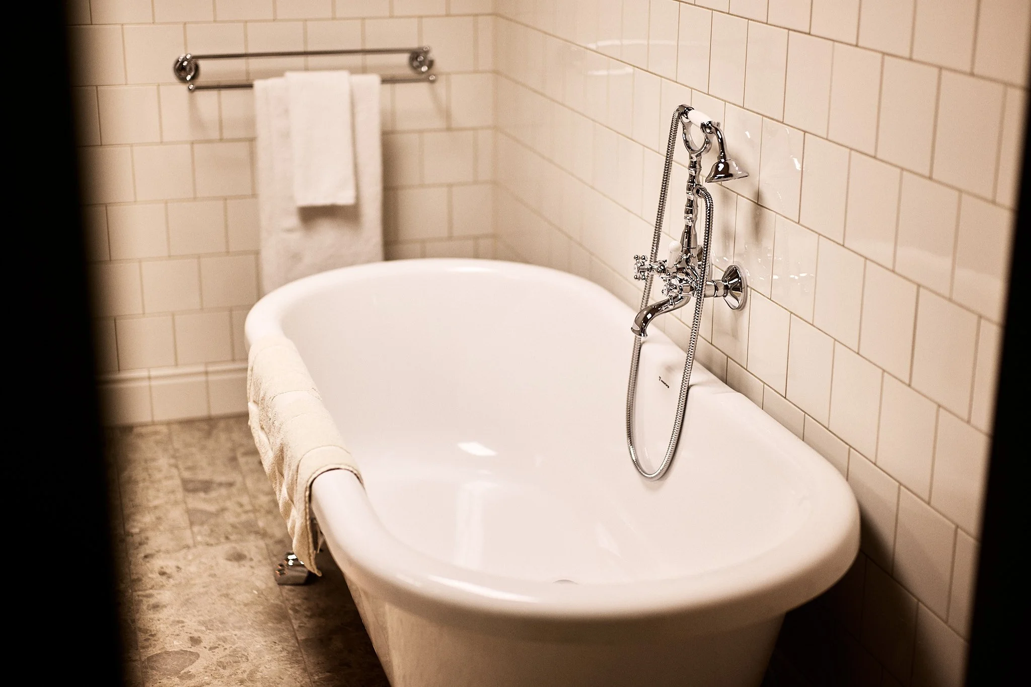 A bathroom with a vintage-style clawfoot bathtub, chrome fixtures, a handheld shower, and a towel hanging on the side, tiled walls, and a tiled floor.