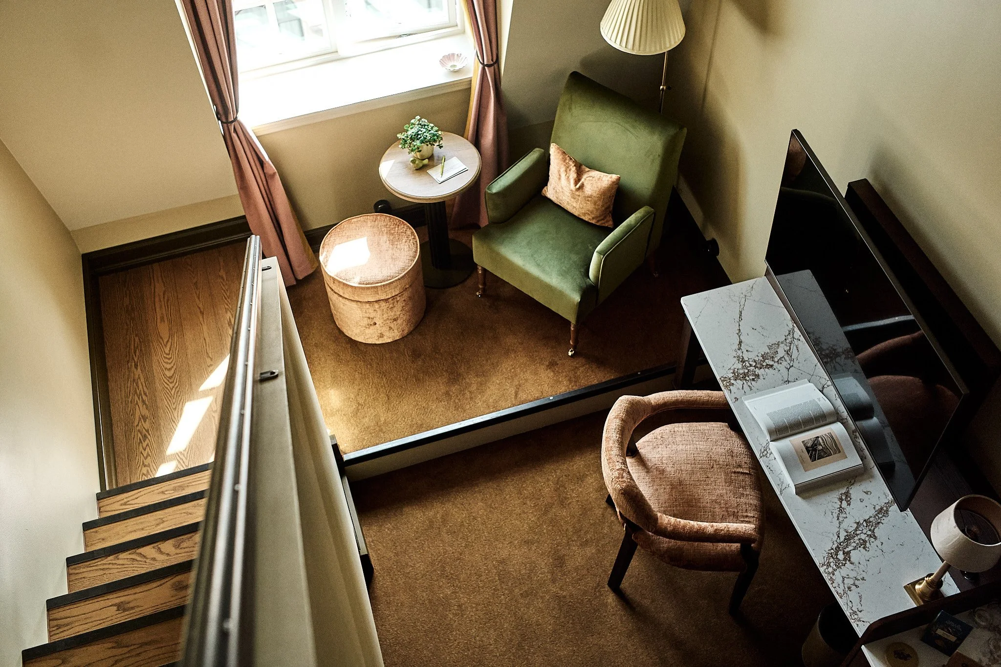 An overhead view of a cozy corner with a green armchair, side table, and floor lamp next to a window with pink curtains. There is a small plant, a notebook, and a pen on the side table, with a round beige ottoman nearby. A TV and a marble-topped furniture piece are also visible.