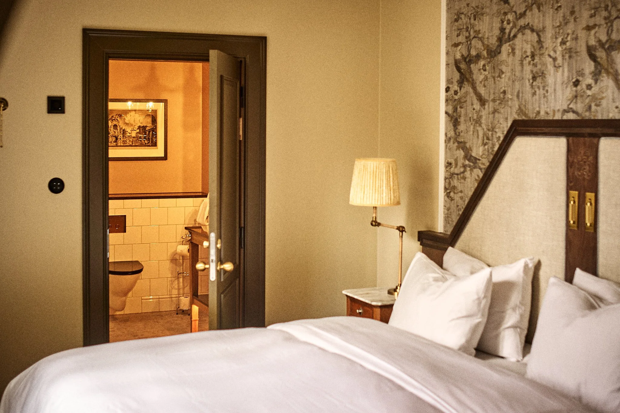 Hotel room with a bed, bedside table, lamp, and view into a bathroom with a toilet and framed artwork on the wall.