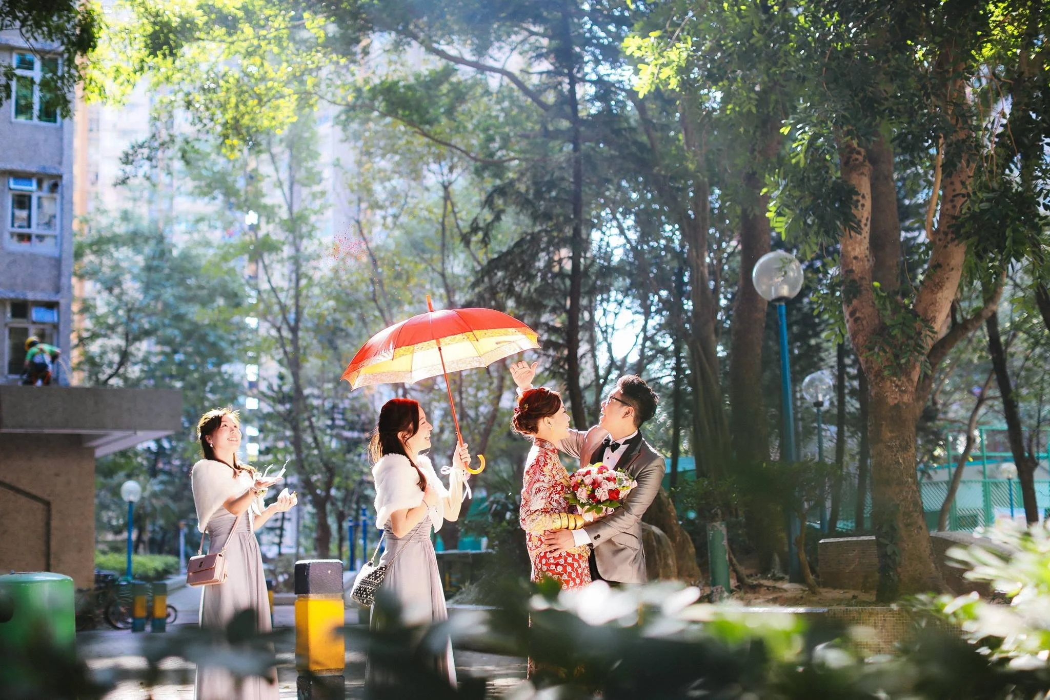 A wedding couple in formal attire is outdoors in a park, with the bride in a red patterned dress and the groom in a gray suit, holding a bouquet of flowers. Two women are standing nearby, one holding a red and yellow umbrella over the couple, and a t