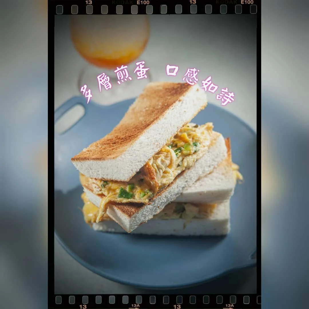Two sandwiches with scrambled eggs on white bread on a blue plate, with a glass of orange juice in the background, and Chinese characters overlaid on the image.