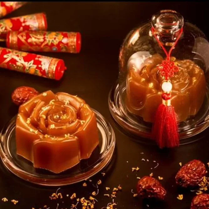 Two rose-shaped caramel desserts on glass plates, decorated with gold flakes, with decorative red and gold tubes and dried fruits on a black surface.