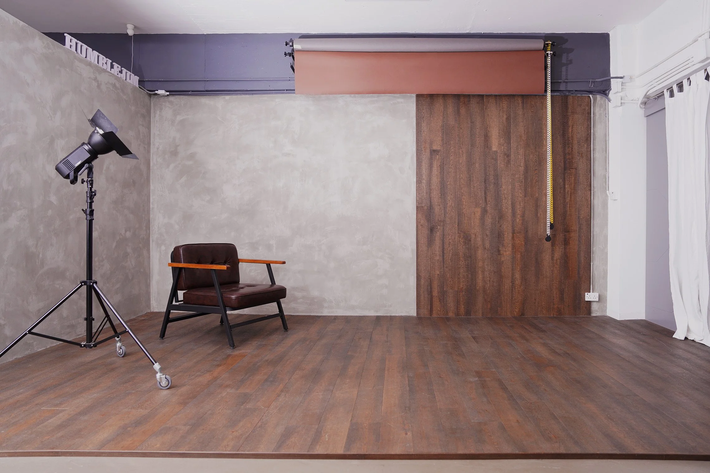 Photography studio with a camera on a tripod, a brown leather chair with wooden arms, a backdrop curtain, and a measurement tape hanging from the ceiling, with a wooden floor and textured wall.