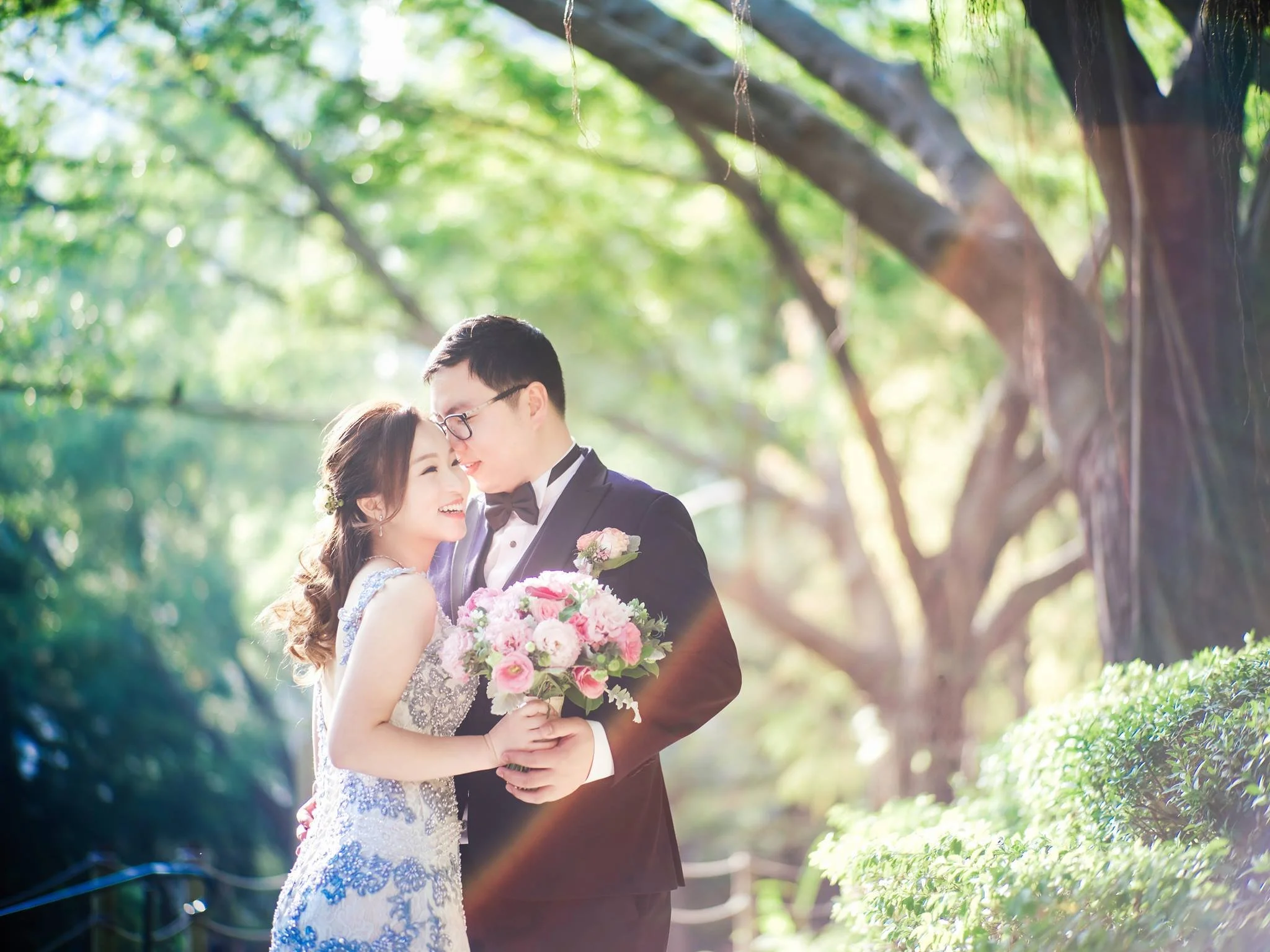 A couple in wedding attire holding a bouquet of pink and white flowers, standing close and smiling in a lush, green outdoor setting.